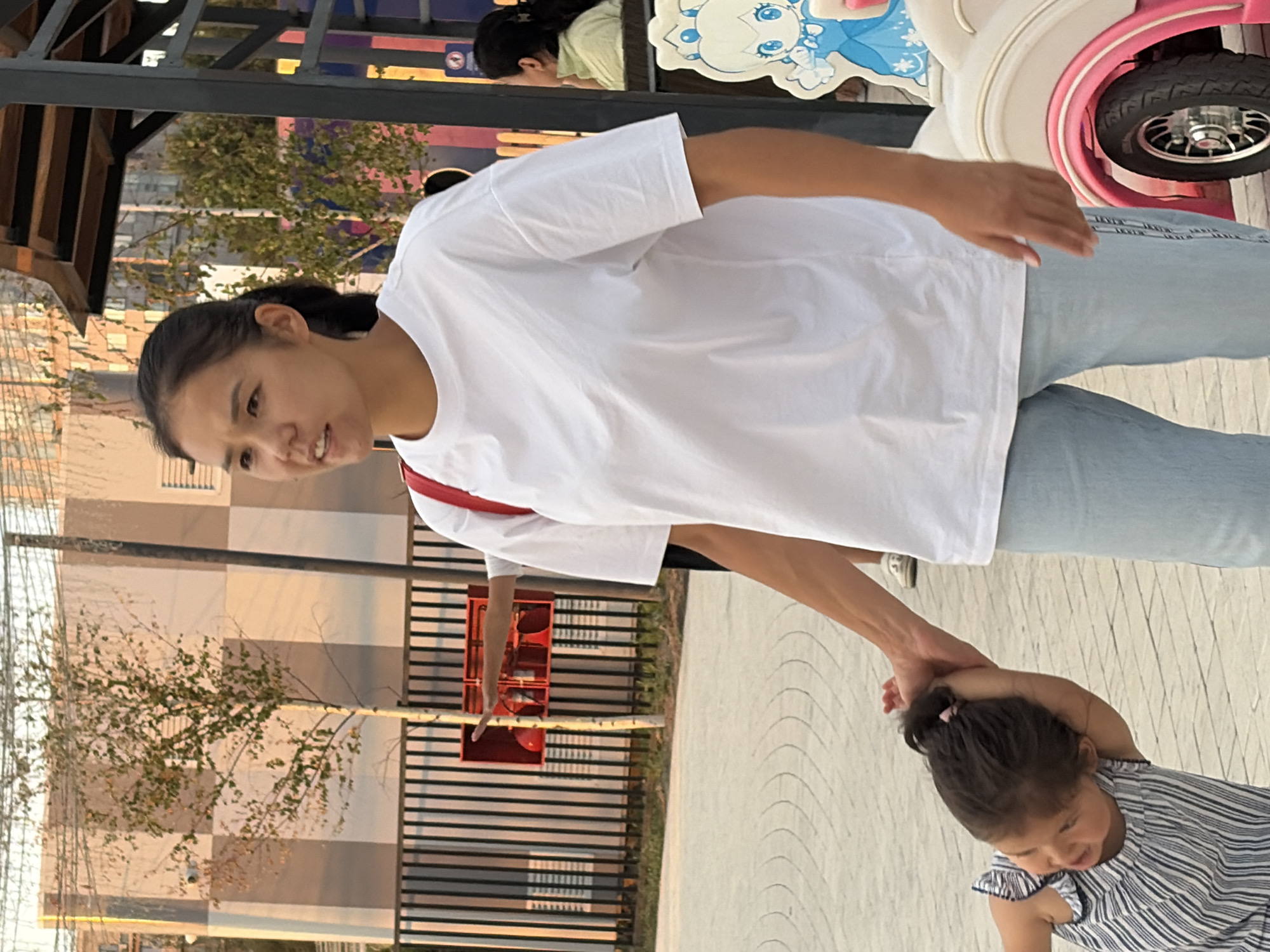 A woman in a white t-shirt and light blue jeans is walking with a young girl in a striped dress. The woman is holding the girl's hand. The girl is looking at the woman and smiling. The scene appears to be outdoors, possibly in a park or a public space. There is a fence in the background, as well as some trees and buildings. A pink and white toy car is visible on the right side of the image.