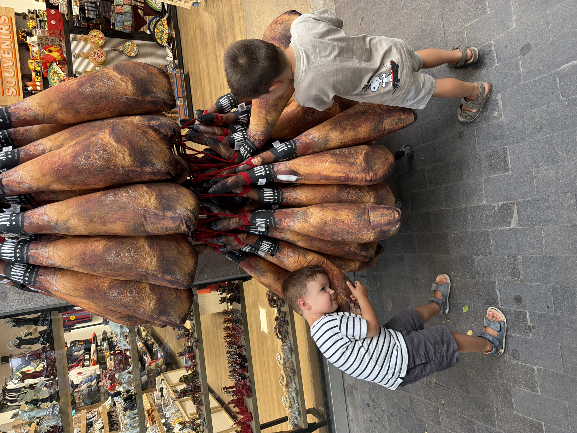 A young boy in a striped t-shirt and grey shorts leans his head against a large cured ham. He wears grey sandals. To the right, another young boy in a grey t-shirt and grey shorts, featuring a Snoopy graphic on the back, looks at the hams. He also wears grey sandals. Both boys are standing on a grey tiled sidewalk. In the background, shelves display various souvenirs and merchandise in a shop.