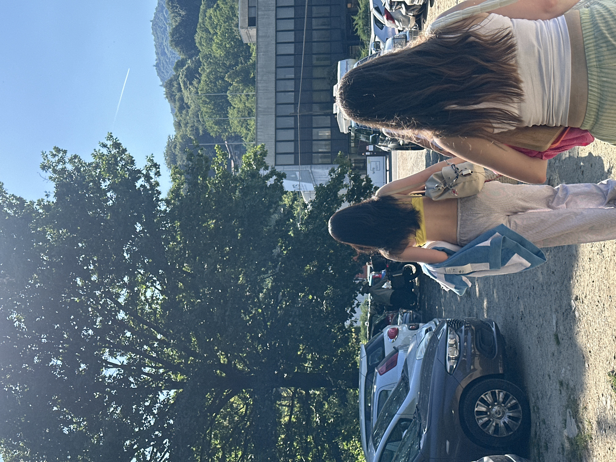 Two women stand facing away from the camera in an outdoor parking area. One wears a white top and striped pants with a pink towel over their shoulder. The other is wearing a yellow top and striped pants with a blue and white towel. Cars are parked on either side of them. In the background are trees, mountains, a building and a blue sky with a contrail. The location is likely San Maurizio d'Opaglio, Italy, based on the surroundings and objects in the image.