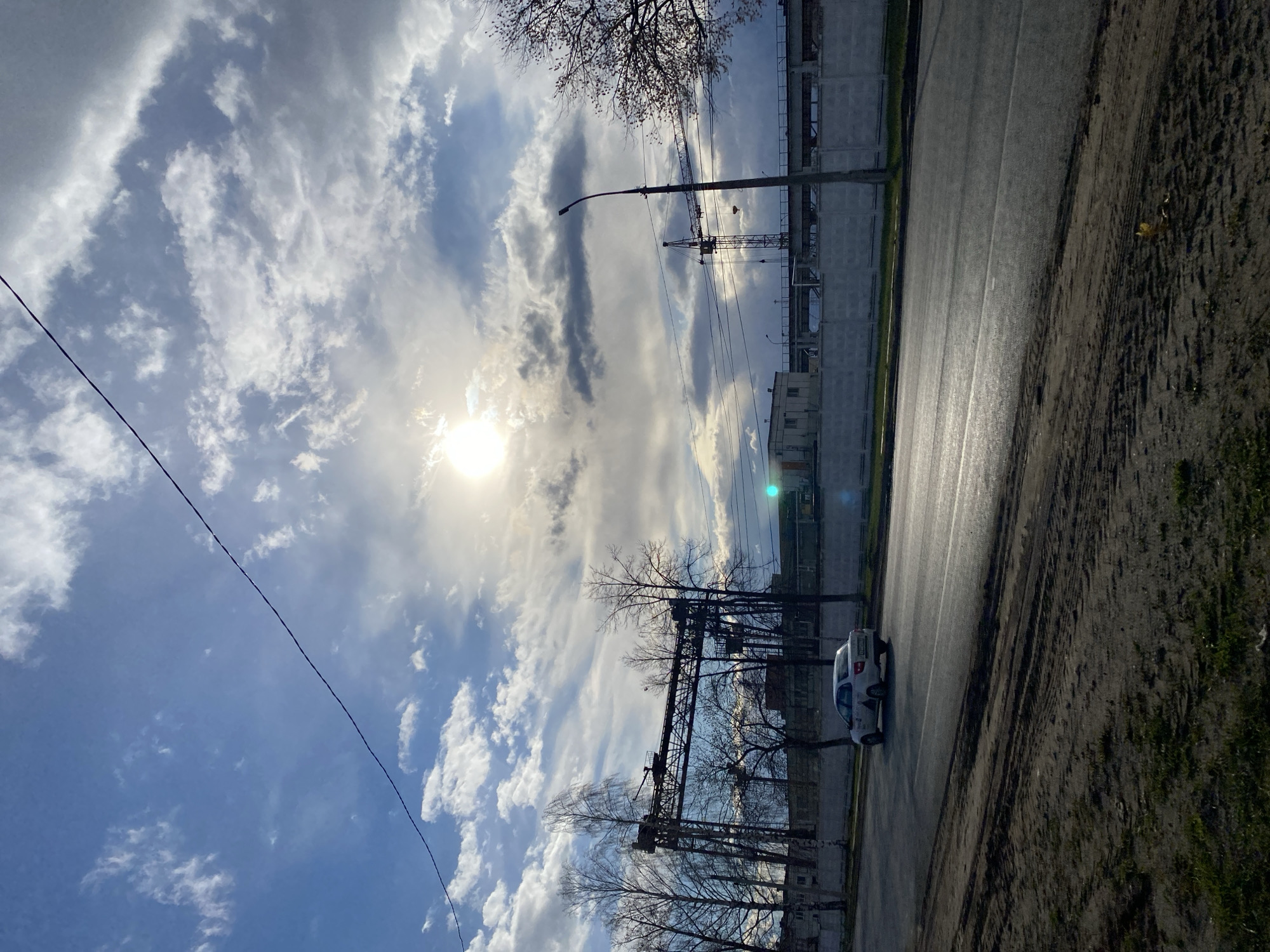 The image displays an outdoor urban-industrial scene in Chaikovsky, Russia, captured on a bright, partly cloudy day. The sky dominates the upper portion, featuring a mix of blue patches and various cloud formations, from wispy cirrus-like streaks to denser cumulus clouds. The sun shines intensely from the upper left, partially obscured by clouds, creating a strong sunburst and lens flare. A dark utility line stretches diagonally across the upper sky.
In the mid-ground and foreground, a road runs from the center towards the background on the right side of the frame. It appears damp and reflective, possibly from recent rain, with a faint white dividing line. To the immediate right of the road is a strip of dark, bare earth marked with tire tracks, leading to a patch of sparse green grass at the far right edge.
On the left side of the road, and extending across the mid-ground, are several bare deciduous trees, their branches devoid of leaves, suggesting late autumn or early spring. Behind these trees, and further to the left, stand large, dark metallic industrial structures, possibly gantry cranes or parts of a factory, backed by concrete walls. A tall lamppost with utility wires is also visible in the mid-ground on the left.
A small, light-colored vehicle, resembling a white hatchback or van, is situated on the road in the mid-ground, facing away from the viewer. Faint, dark markings or text are visible on its side, but are illegible. The scene is static, without any discernible human activity. The overall impression is that of a quiet industrial or semi-urban periphery.