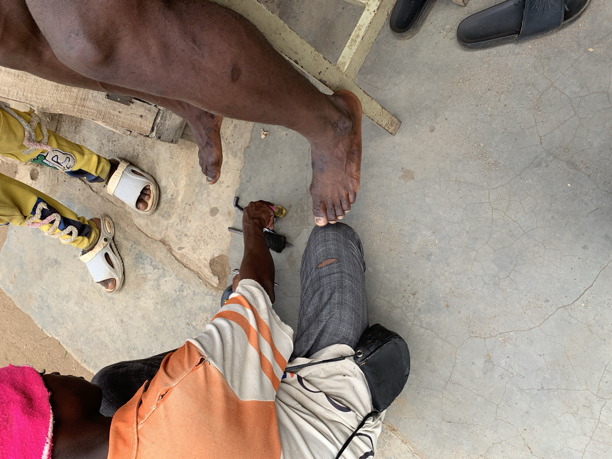 The image shows a view from above of three individuals on a cracked concrete surface in Rafin Par, Nigeria. One individual is sitting and appears to be grooming the toenails of another individual, who is standing. A small child is seated nearby wearing yellow pants and white sandals. The groomer is wearing gray pants and a white t-shirt with orange stripes. A pink towel is on their head. The person having their nails trimmed is dark-skinned. The groomer holds a tool, possibly a clipper or nail file, in one hand and appears to be working on a toe. A pair of black sandals are in the upper right corner of the image. A metal frame is partially visible in the image.