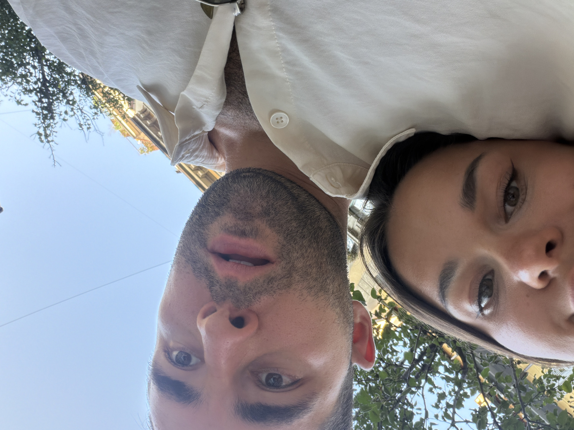 A close-up selfie shows a man and a woman looking up towards the camera. The man's face, mostly in focus, is positioned in the right half of the frame, with his mouth slightly open and his eyes looking directly at the camera. He has short, dark hair and stubble. The woman's face, slightly out of focus, rests on the man's shoulder, her eyes also looking towards the camera. She has dark hair and is wearing a light-colored shirt. The background consists of a clear blue sky, some trees with green leaves, and buildings that appear to be in an urban setting. The overall lighting suggests it is daytime. There are no specific landmarks visible that would indicate the city of Milan, Italy.