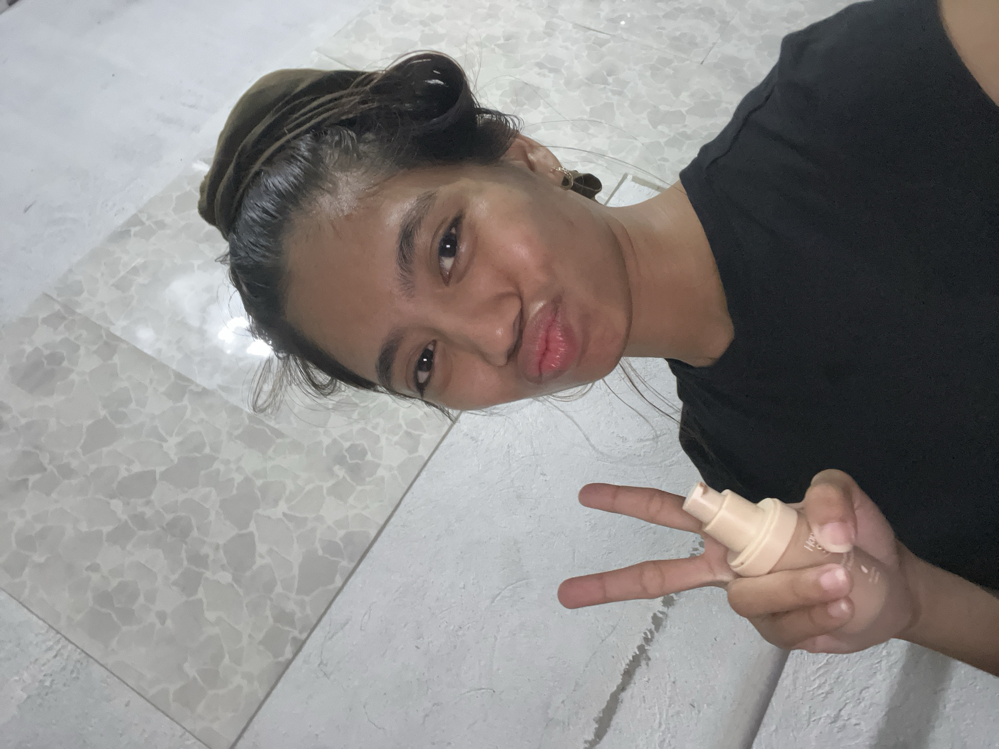 The image features a young individual, likely female, posing playfully for the camera in an indoor setting, consistent with a home in Davao City, Philippines. The image is captured from a slightly overhead and tilted angle, adding to its candid feel.
The subject has dark hair, styled loosely and pulled back, with some strands framing her face. She is wearing a plain black t-shirt. Her expression is playful, characterized by dark eyes looking directly at the viewer and a 'pouty' or 'duck face' with slightly parted, full lips. Her skin appears dewy and moisturized. A small, simple hoop earring is visible in her right ear.
In her right hand, held prominently, is a light beige-colored cosmetic bottle with a pump dispenser, likely a foundation or similar liquid makeup product. Her index and middle fingers are extended in a peace sign gesture, holding the bottle between them. A small amount of product residue is visible on her fingertips.
The background consists of light-toned, possibly white and gray, tiled surfaces with an irregular, speckled or mosaic-like pattern. This tiled surface appears to be a floor or a low wall. The lighting is soft and even, typical of an indoor environment, but does not provide clues about the time of day. There is no visible text in the image. The overall emotion conveyed is one of casual playfulness and self-assurance.