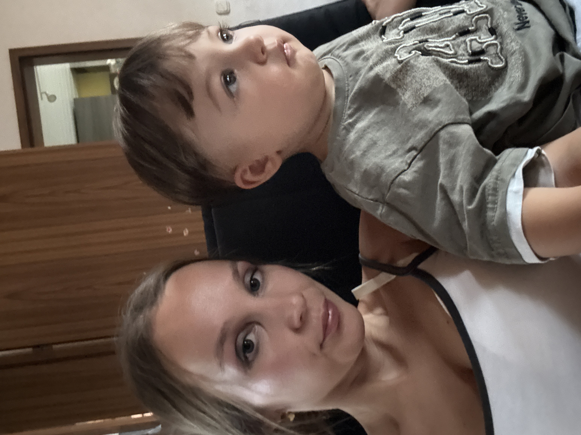 A woman and a young child are seated next to each other indoors. The woman is on the left, facing the camera with a neutral expression. She has blonde hair pulled back and is wearing a white tank top with a dark trim. The child is on the right, looking off to the right side of the frame. The child has short brown hair and is wearing a grey t-shirt with a graphic design. The background shows a wooden door and a wall, suggesting an indoor domestic setting. The scene appears to be in Überherrn, Germany.