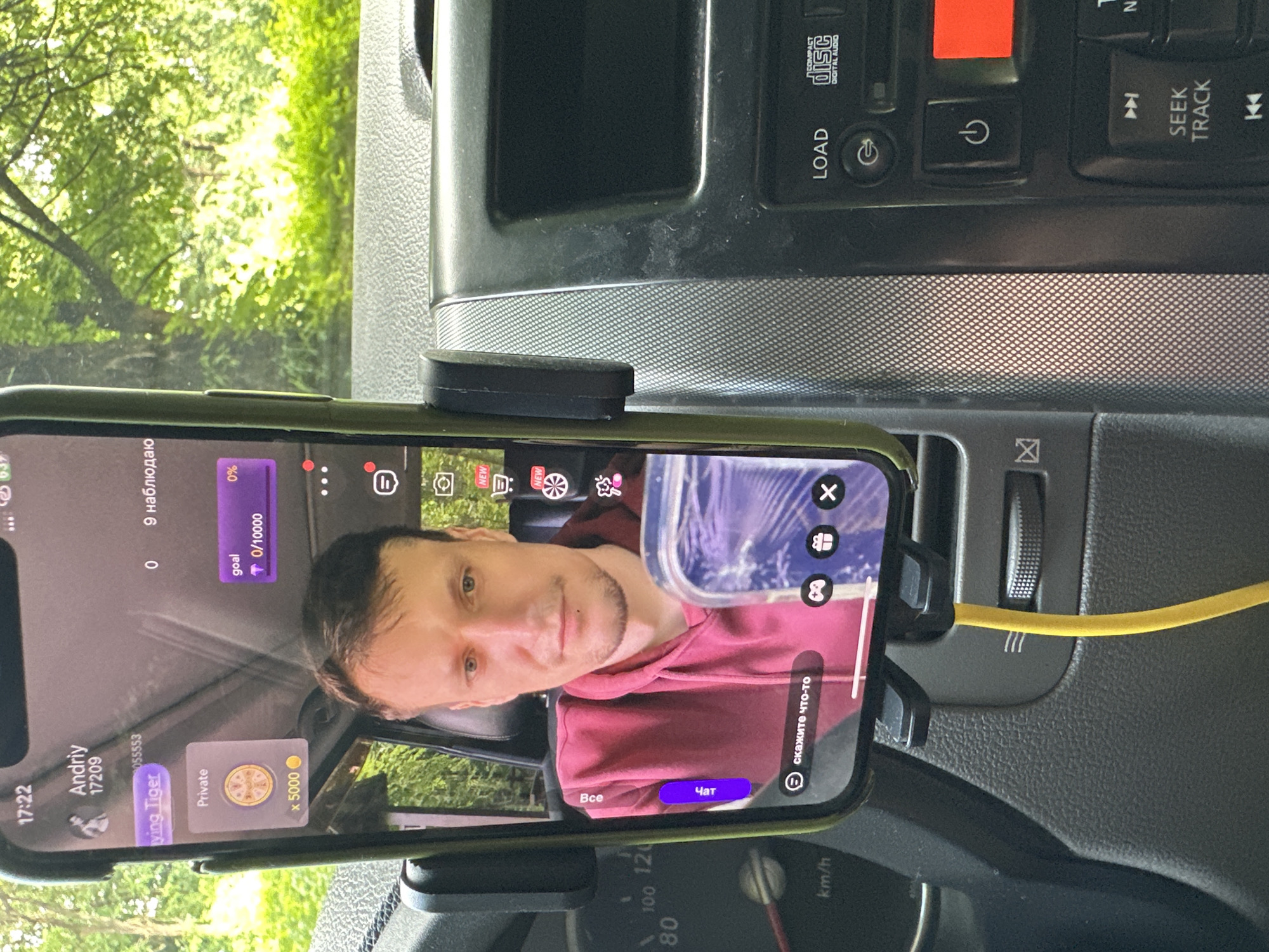 The image shows a person inside a vehicle, with a smartphone mounted on the dashboard. The smartphone screen displays a live streaming application, showing the face of the person, identified as "Andriy", along with various streaming interface elements such as "goal" progress, "Private" chat options, and a username "Flying Tiger". The vehicle's dashboard is visible, with a speedometer and other controls. The background through the windshield is out of focus, showing green foliage and trees, suggesting an outdoor setting. The context of Shatava, Ukraine is not directly derivable from the visual information present in the image.