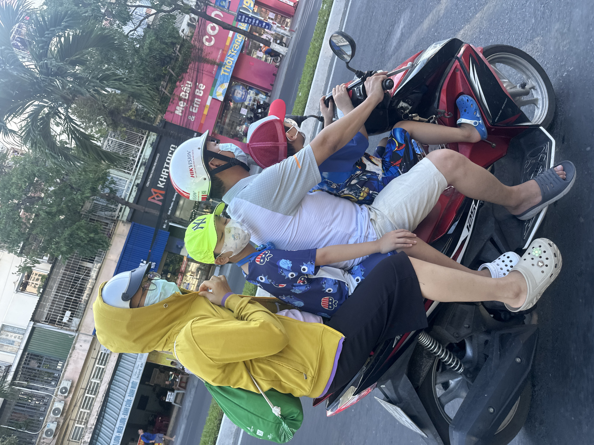 A family of four, including two adults and two children, are riding on a red and black motorcycle through Bac Nha Trang Ward, Vietnam. The driver, a man wearing a white helmet with red accents and a gray shirt, is in the front. The woman behind him is wearing a bright yellow hooded jacket and a light blue face mask. In front of her, a young boy in a blue shirt with cartoon characters and a neon green cap is also wearing a face mask. Another child, wearing a red and white hat and a face mask, is seated behind the boy. The children are wearing casual clothing and sandals, and the woman has on black pants and light beige clogs with green embellishments. The motorcycle is in motion on a paved road with buildings and trees visible in the background. The buildings appear to be commercial establishments, with signs in Vietnamese. The scene suggests a typical urban environment in Vietnam.