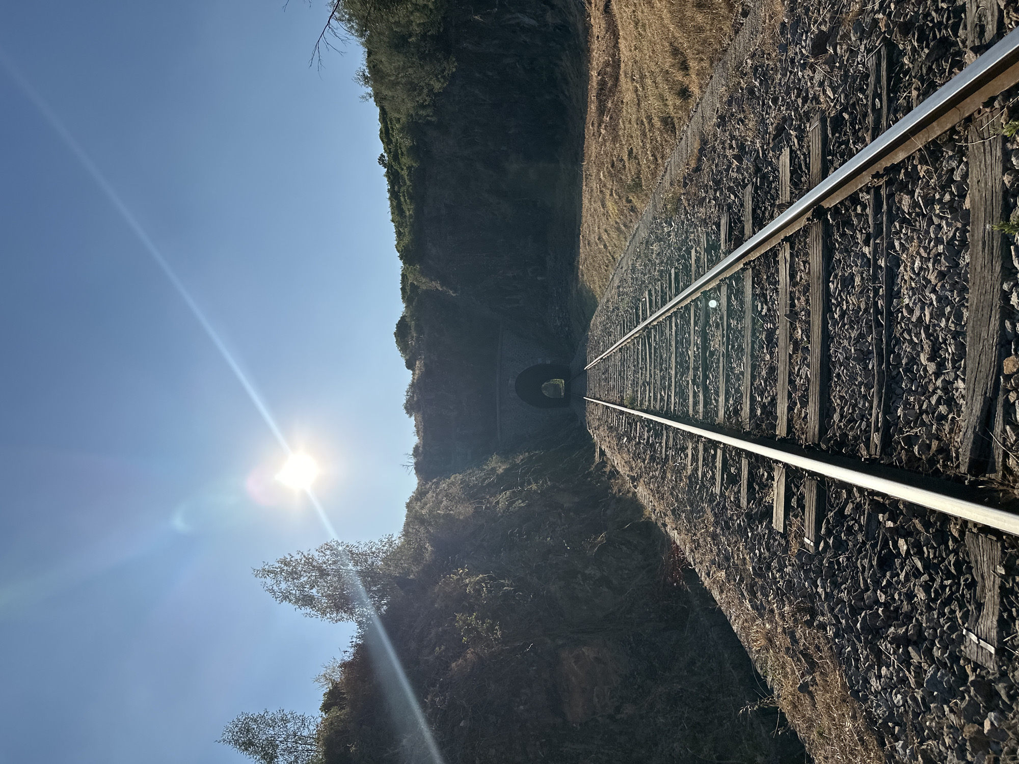This is a straight-on, eye-level, medium shot of a set of railway tracks leading towards a tunnel entrance. The sun is shining brightly in the upper left quadrant of the sky, casting lens flares across the image. The tracks are made of metal rails and wooden sleepers, laid on a bed of gravel. The tracks are flanked by dry, grassy terrain on the right and a steep, rocky embankment covered in vegetation on the left. The tunnel entrance is visible in the distance, a dark, arched opening set into a hillside covered with trees and bushes. The sky is clear and blue. The image was taken outdoors on a sunny day. The location appears to be rural.