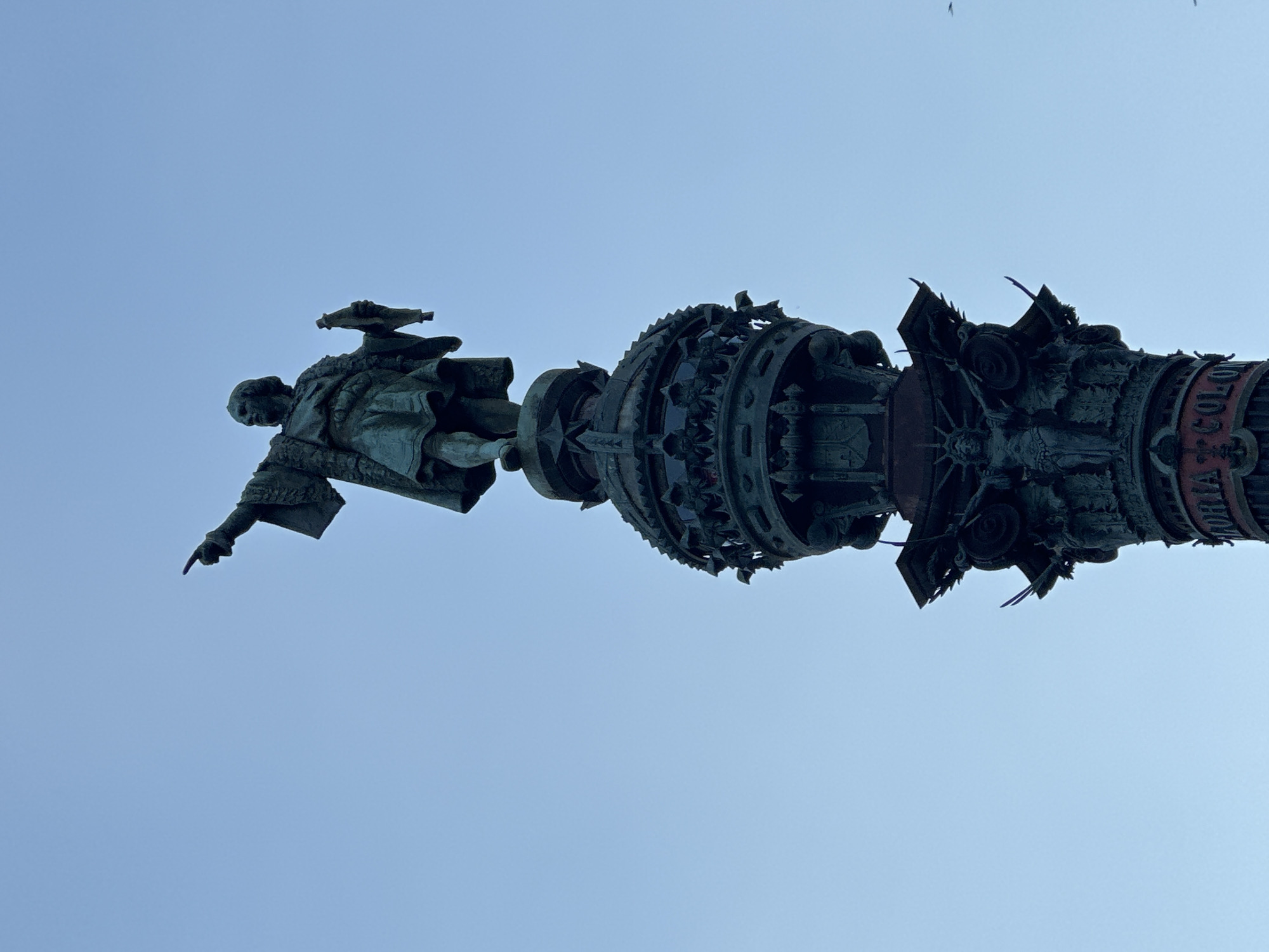 The image shows the Monument to Columbus in Barcelona, Spain, against a clear, pale blue sky. The monument is a tall, ornate column topped with a statue of Christopher Columbus. The statue depicts Columbus in a historical costume, with one arm extended and pointing forward, and the other holding a scroll. The column itself is richly decorated with detailed carvings and architectural elements, including what appears to be reliefs and decorative motifs. The overall color palette is dominated by the dark, weathered bronze of the monument and the soft blue of the sky. The perspective is from below, looking upwards at the monument. A few small birds are visible in the upper right quadrant of the image. The base of the monument, partially visible at the bottom, has some lettering that appears to read "ORIA COLON".