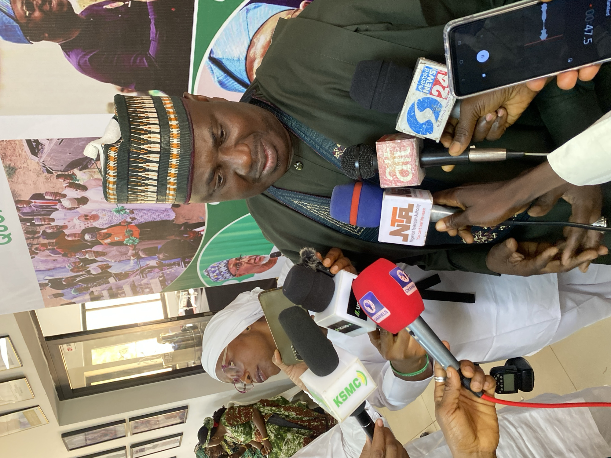 An indoor scene captures a media engagement in Kwarau, Nigeria, featuring a man addressing a group of reporters. The central figure, dressed in an olive-green traditional tunic and a patterned cap, is speaking into multiple microphones. His expression is calm, with a slight smile.
Numerous hands, belonging to various media personnel, are visible extending microphones and recording devices towards the speaker. Prominently displayed microphone logos include "KSMC," "CHANNELS" (on a red microphone), "silverbird NEWS 24," and "NTA" (Nigerian Television Authority), the latter bearing the tagline "You can't beat the reach." One individual is seen holding a smartphone actively recording audio, with a timer showing "00:47.5" and a pause icon on its screen. Another person in a white head covering and glasses is also holding a microphone, suggesting their involvement as a reporter or participant.
In the background, a large green and white banner is partially visible, featuring blurred images of people, some wearing traditional attire, and partial text including "QU." Below the banner, framed pictures adorn a light-colored wall, and a window reflecting bright light is visible further back. The overall setting appears to be a formal press conference or interview session within a building, likely a media center or public office. The artificial lighting or bright window light suggests the event could be taking place during daytime.