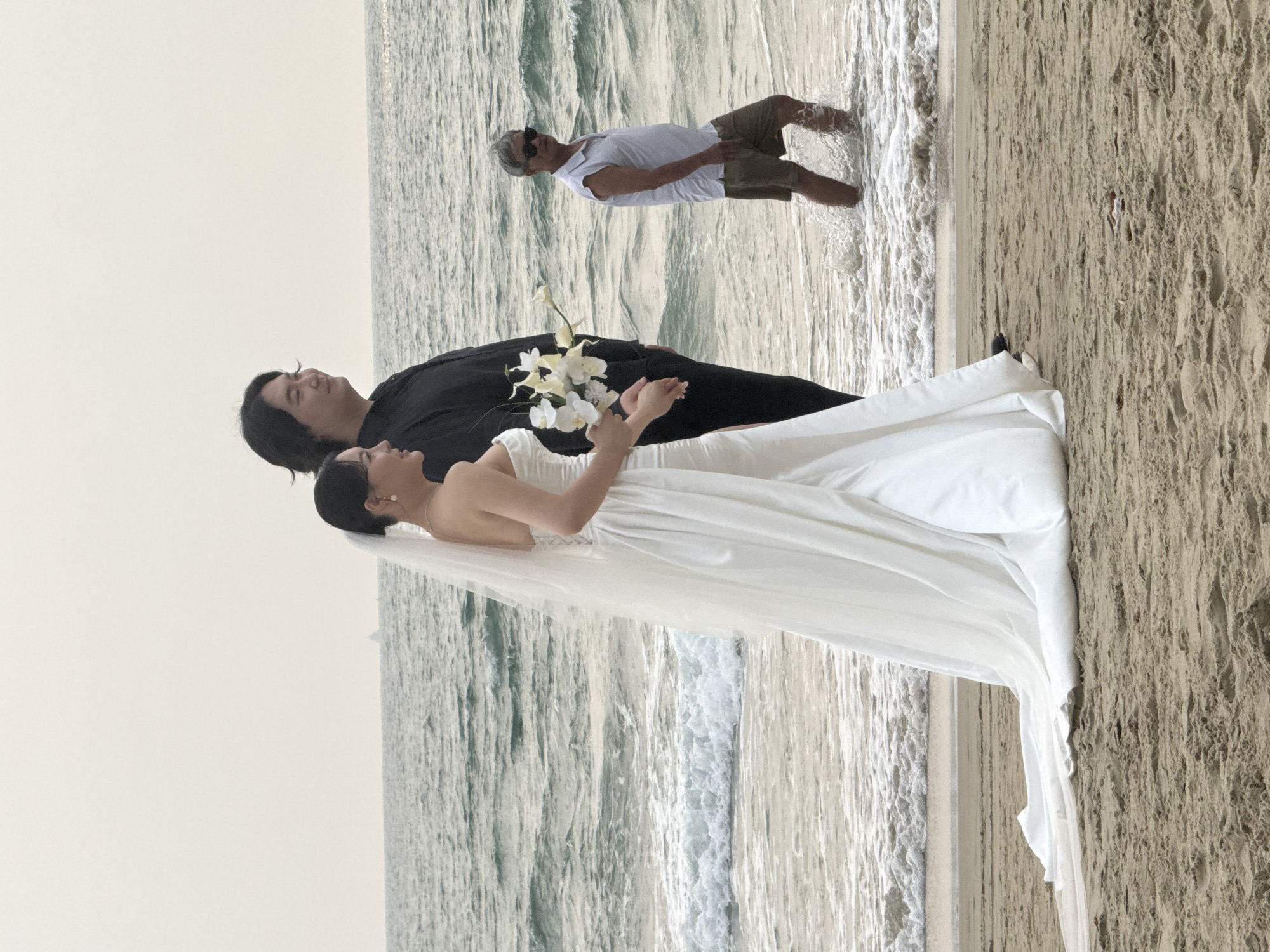 A wedding photo is taken on a beach. A bride in a white wedding dress holds a bouquet of white flowers and stands next to a groom in a black shirt. They are standing near the water. Behind them, an older man in a white t-shirt and tan shorts stands in the water. The location is likely in Đà Nẵng, Vietnam, given the coastal environment. The sand on the beach is a light brown color. The sky is overcast.