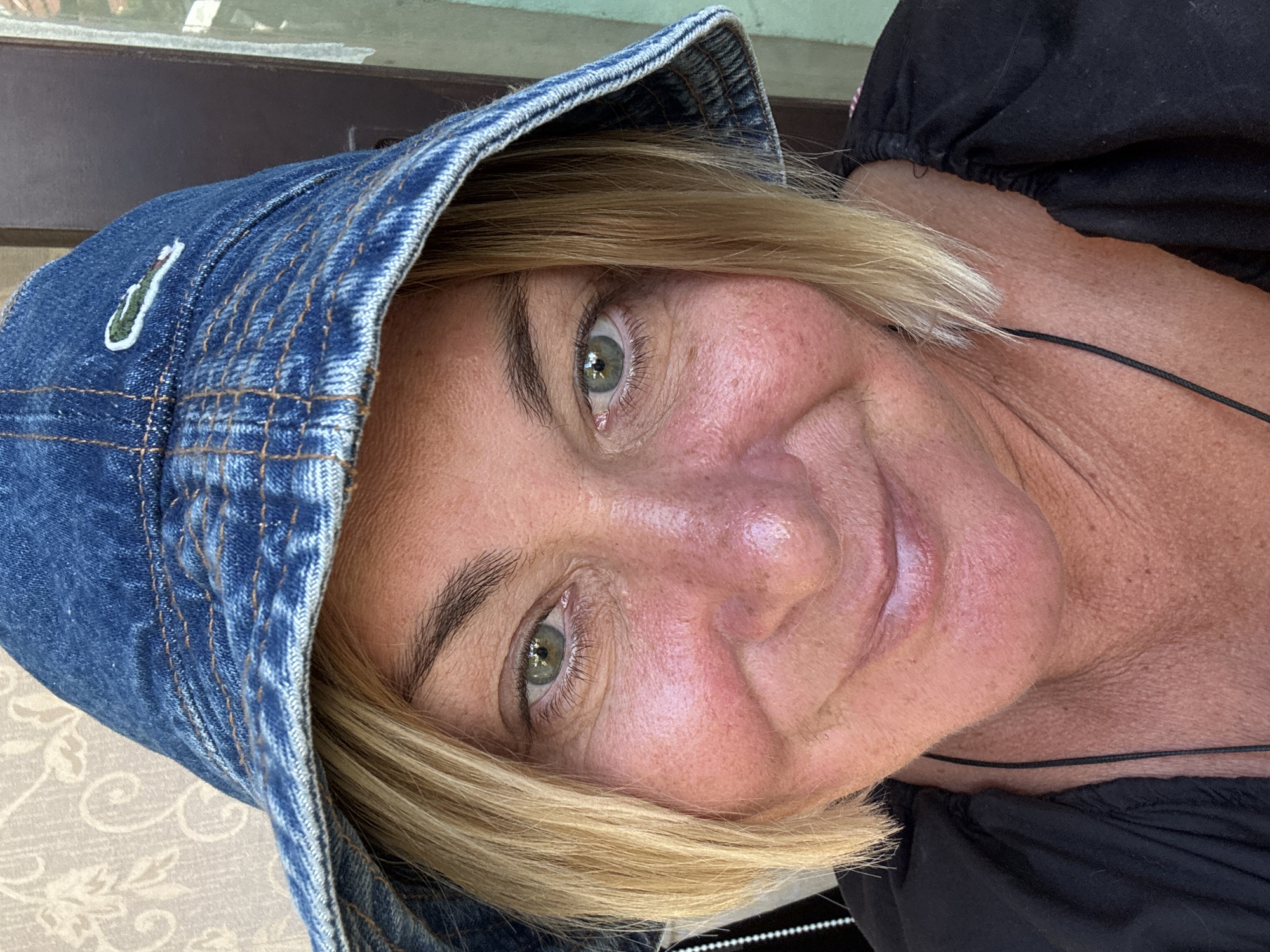 A woman is depicted in a close-up, angled portrait wearing a denim bucket hat with a Lacoste logo. Her skin is fair and shows signs of aging, particularly around the eyes and cheeks. She has light blonde hair framing her face and green eyes. She is wearing a black garment. The background appears to be an outdoor or semi-outdoor setting with muted colors. There's no evidence to support the location is in the city of บ้านบางรักษ์, Thailand.