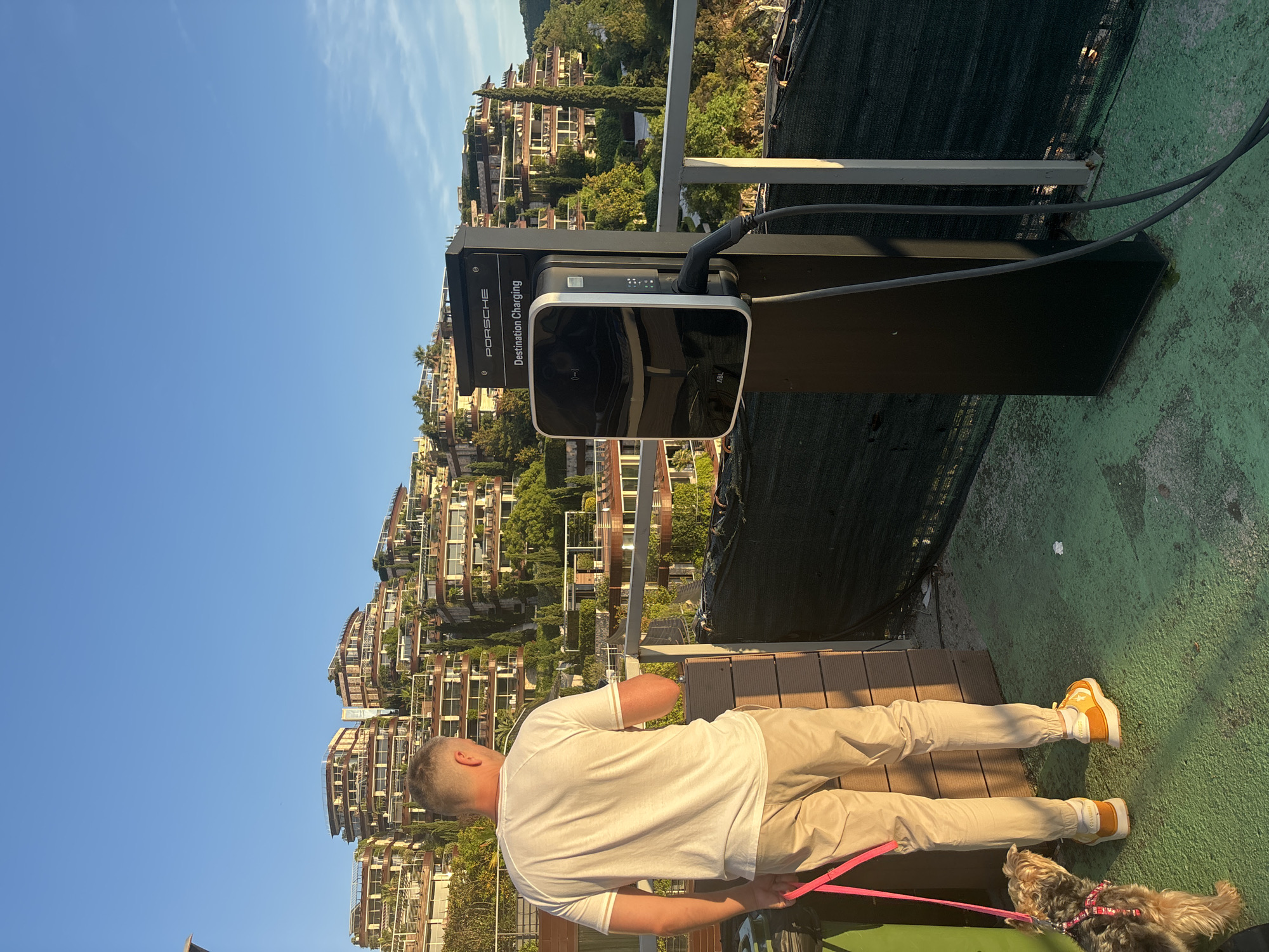 The image captures a man and his dog standing near an electric vehicle charging station, overlooking a picturesque hillside dotted with buildings under a clear blue sky. The scene is set at a high vantage point, likely a terrace or balcony, with a green-colored ground surface and a black safety net or fence on the right.
In the foreground, a man, seen from behind, is wearing a white short-sleeved t-shirt and light beige or khaki pants. He has short hair and is holding a pink leash. Beside his feet is a small, fluffy dog, possibly a Yorkshire Terrier, also on the pink leash. The man is wearing yellow and white high-top sneakers.
To the right of the man stands a black Porsche Destination Charging station, which appears to be actively in use with a charging cable plugged into the unit. The text "PORSCHE" and "Destination Charging" are prominently displayed on the station. The charging unit itself also shows the brand name "ABL".
The background reveals a stunning landscape of a sun-drenched hillside, densely covered with multi-story residential or resort buildings characterized by terraces and lush green foliage, suggesting a Mediterranean-style architecture. This aligns with the provided context of Budva, Montenegro. The sky is bright blue with only a few wispy clouds, indicating clear weather and a sunny day, likely late morning or afternoon given the strong lighting and shadows.