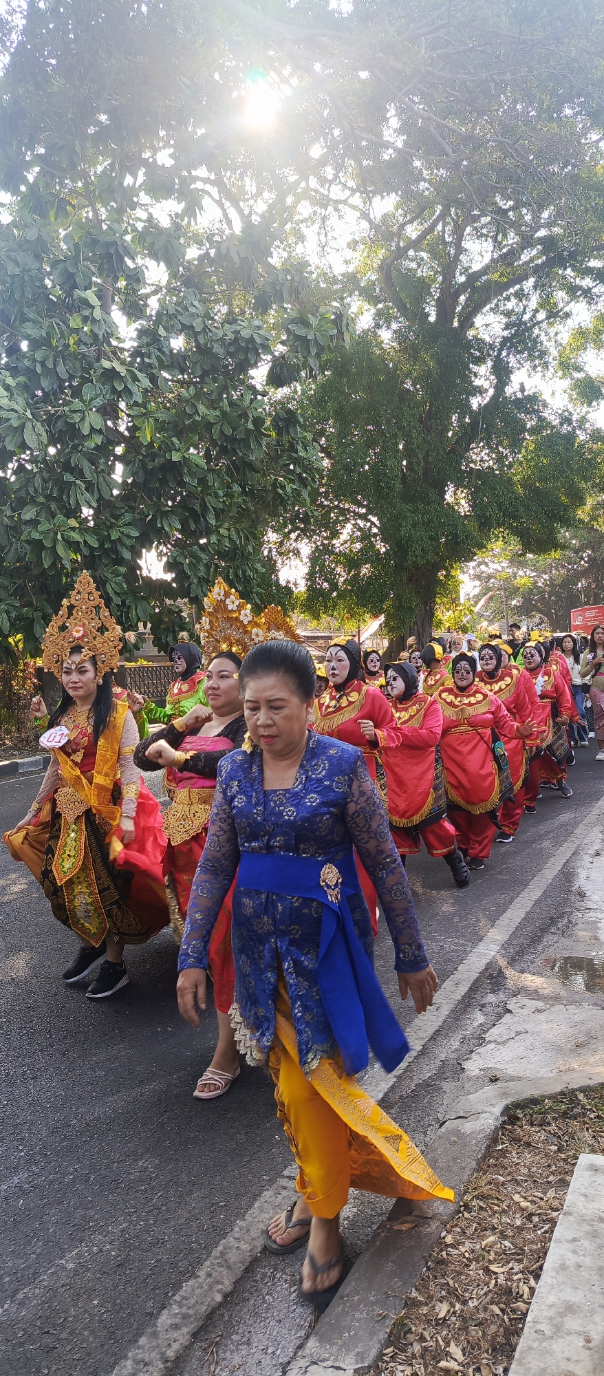 A group of women in traditional Indonesian attire are walking in a procession down a street in the city of Malang, Indonesia. The woman in the foreground is wearing a blue lace top with gold accents and a yellow sarong. She is walking with a blue sash around her waist. Behind her, other women are dressed in vibrant red and green costumes, some with face paint. They are adorned with ornate gold headdresses and jewelry, suggesting a cultural performance or festival. The procession is taking place on a paved street with a concrete curb on the right side. Lush green trees line the street, and the sun is shining brightly, creating lens flare in the upper portion of the image. The overall scene depicts a lively street event with participants dressed in elaborate, culturally significant clothing.