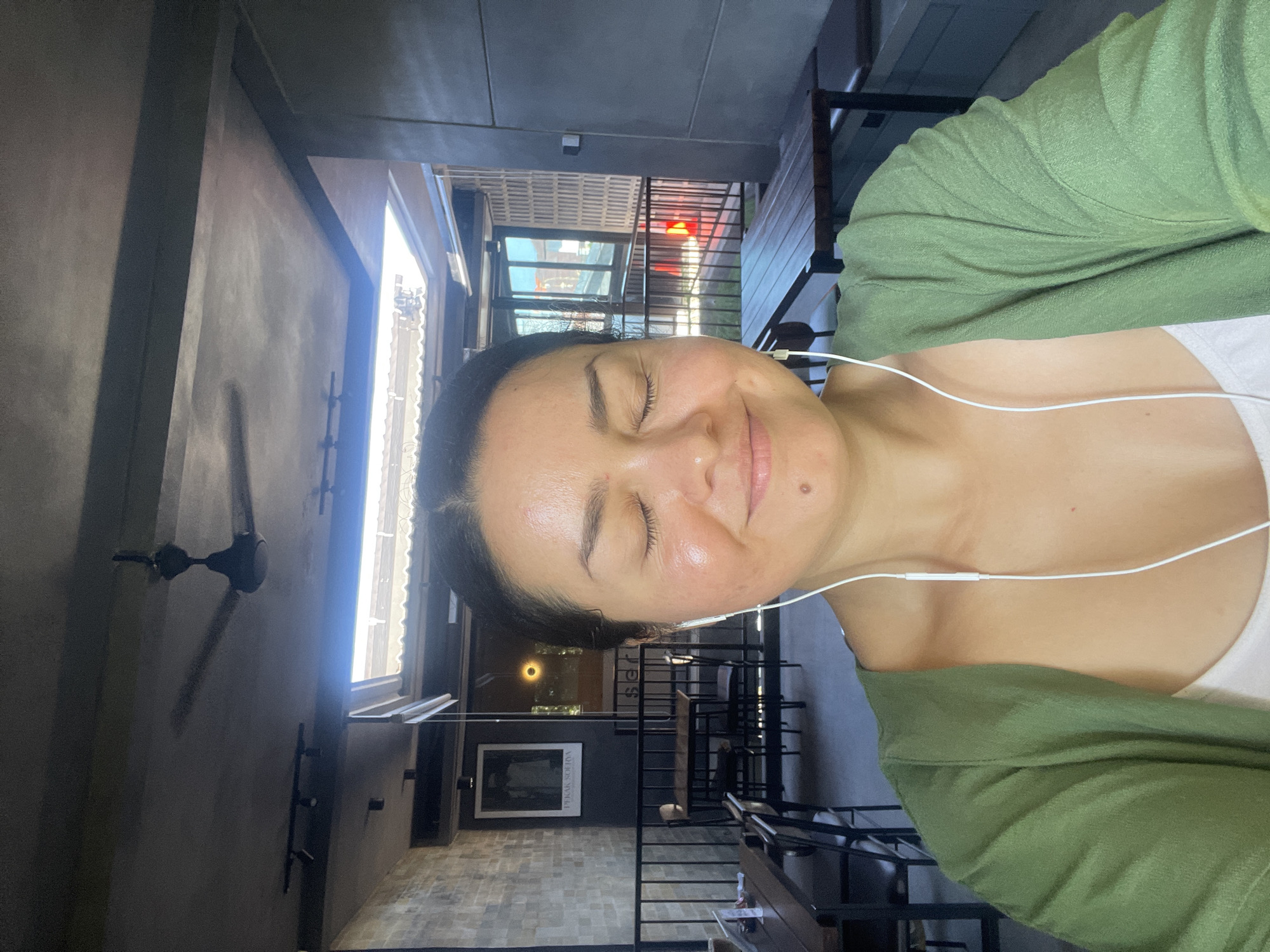 A medium shot photograph captures a woman with her eyes closed, wearing white earbuds and a green cardigan over a white top. She is situated in what appears to be an indoor seating area, possibly a cafe or restaurant, with a ceiling fan visible overhead. The background reveals tables and chairs, a brick wall, and windows that allow natural light to enter. The setting suggests an urban environment, potentially in Kuta Selatan, Indonesia, given the lighting conditions and architectural elements common in the region. The woman's pose implies a moment of relaxation or listening to music.