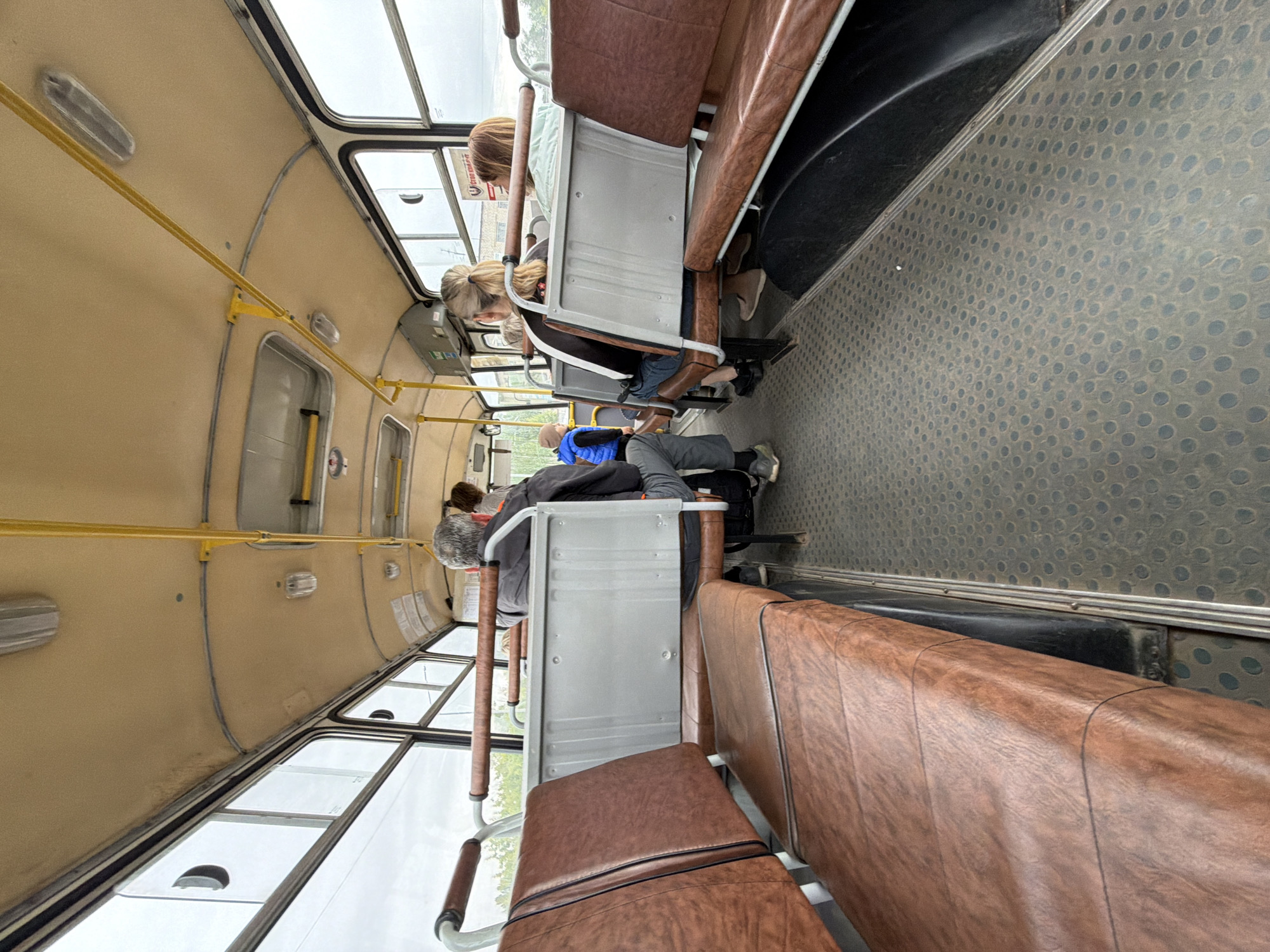 The image shows the interior of a bus, likely in Chaikovsky, Russia, as indicated by the location context. The bus has rows of brown leatherette seats with metal frames and yellow grab bars running along the ceiling. The floor is a grey, textured material with a pattern of circular indentations. Large windows line the sides of the bus, offering a view of an overcast sky and some greenery outside.
Several passengers are visible, seated and seemingly in transit. In the foreground, a person's legs are visible, wearing grey trousers and dark shoes. Further back, another passenger is seated with their head resting on their arms. Towards the middle of the bus, a person with blonde hair pulled back in a ponytail is seen looking down. Another passenger with lighter brown hair is seated behind them.
The lighting suggests it might be daytime, with the overcast sky contributing to a diffused light. The overall atmosphere appears calm and ordinary, consistent with daily public transportation. No specific text is clearly legible in the image.