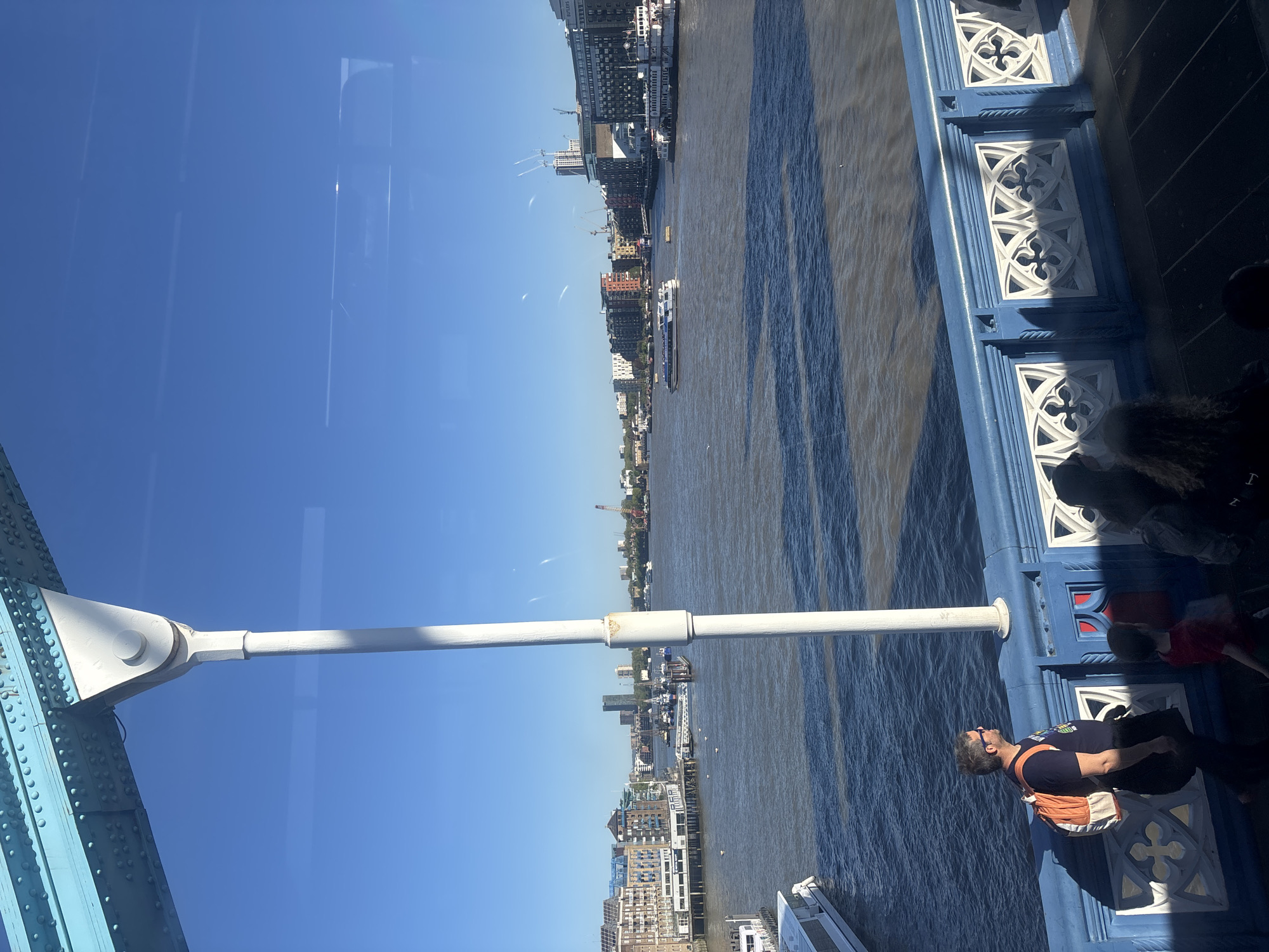 This media file captures an outdoor scene from a high vantage point on a bridge overlooking the River Thames in London, United Kingdom, under clear blue skies. The image is taken through what appears to be a glass barrier, as reflections of the bridge structure and sky are visible.
The prominent subject on the right is a section of the bridge's ornate blue and white railing, featuring distinctive gothic-style quatrefoil designs. Directly behind this railing, partially obscured, are several people, suggesting a sightseeing activity. One individual in the foreground, wearing a dark blue t-shirt, sunglasses, and an orange backpack, is clearly visible, looking out towards the river. A child in a red top is visible further back. A white pole or support beam extends vertically from the bridge structure into the frame.
The central part of the image shows the wide, murky brown expanse of the River Thames. The water's surface reflects the sunlight, creating patterns, and shadows from structures or elements of the bridge are cast upon it. Several boats, including what appears to be a larger passenger vessel and smaller craft, are seen navigating the river.
Along the far bank of the river, a dense cityscape stretches into the distance. A mix of modern buildings, including multi-story residences and office blocks, and older architectural styles are visible. Construction cranes are also discernible in some areas. Faint text, such as "BUTLER'S WHARF," can be identified on one of the buildings, confirming the London context.
The bright sunlight indicates it is daytime, likely midday or early afternoon, with no clouds in the sky, suggesting pleasant weather for outdoor activities. The overall impression is one of urban sightseeing on a clear day from a prominent London landmark, likely Tower Bridge.
