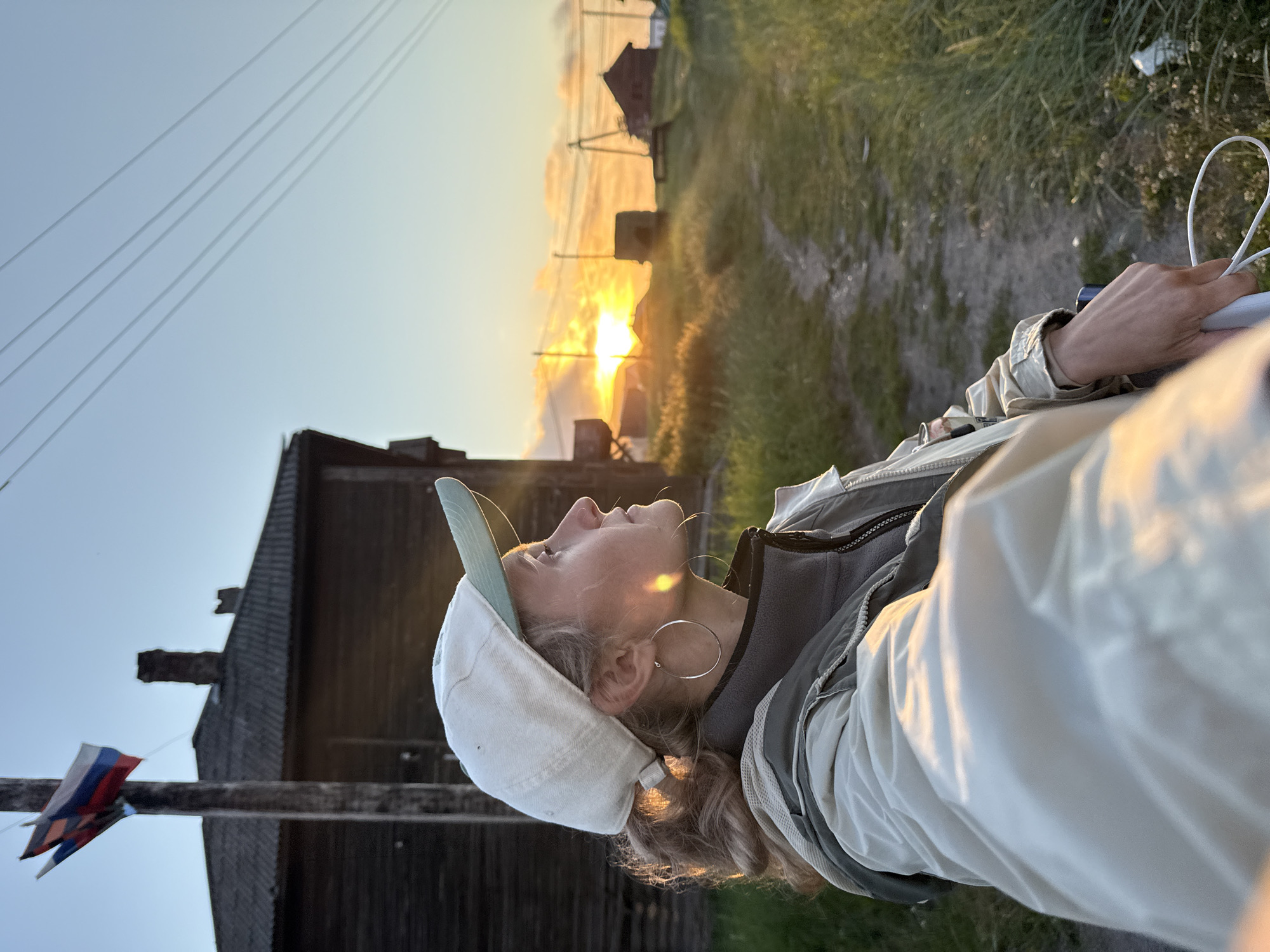 A woman wearing a white baseball cap, a grey fleece jacket, and a beige jacket stands on a dirt path in Teriberka, Russia, at sunset. Two Russian flags fly from a wooden pole behind her. In the background, wooden buildings and power lines are visible against a sky lit by the setting sun. The woman is looking towards the sunset.