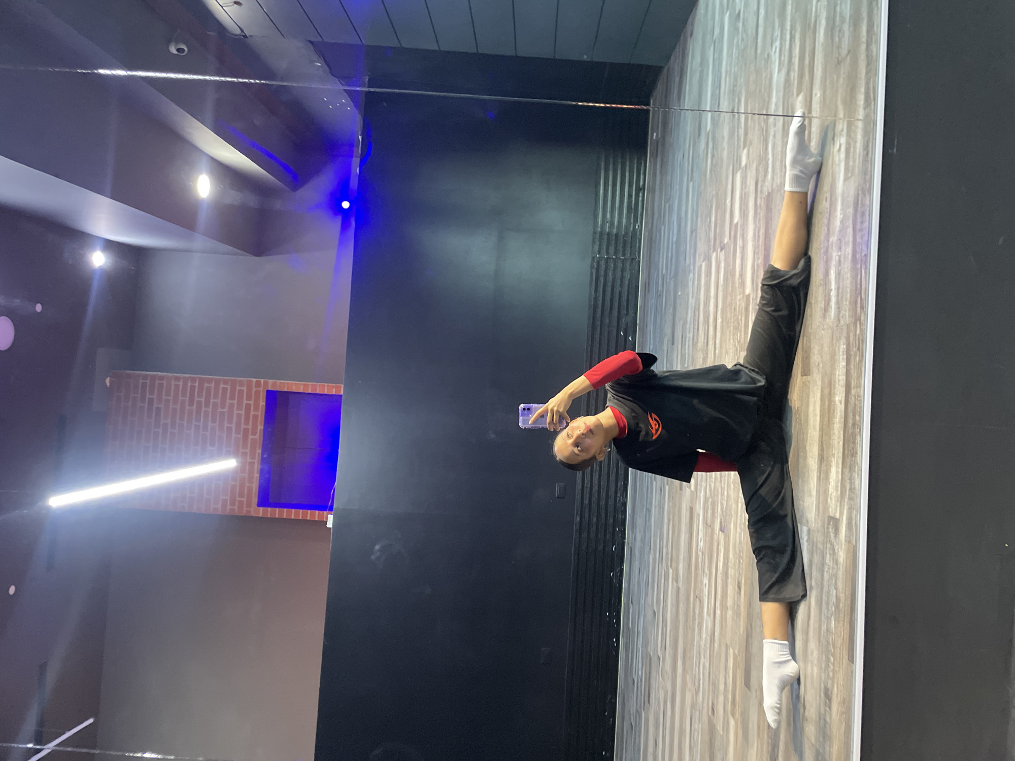 A young woman is performing a split on the floor in a dance studio. She is wearing a black t-shirt with an orange logo, a red long-sleeved shirt underneath, dark pants, and white socks. She is holding a purple smartphone in her right hand, taking a selfie in a large mirror that occupies the right side of the frame. The studio has wooden floors and dark walls, with some decorative brickwork visible in the background. Blue lighting is also present in the background. The setting appears to be a modern dance or fitness studio.