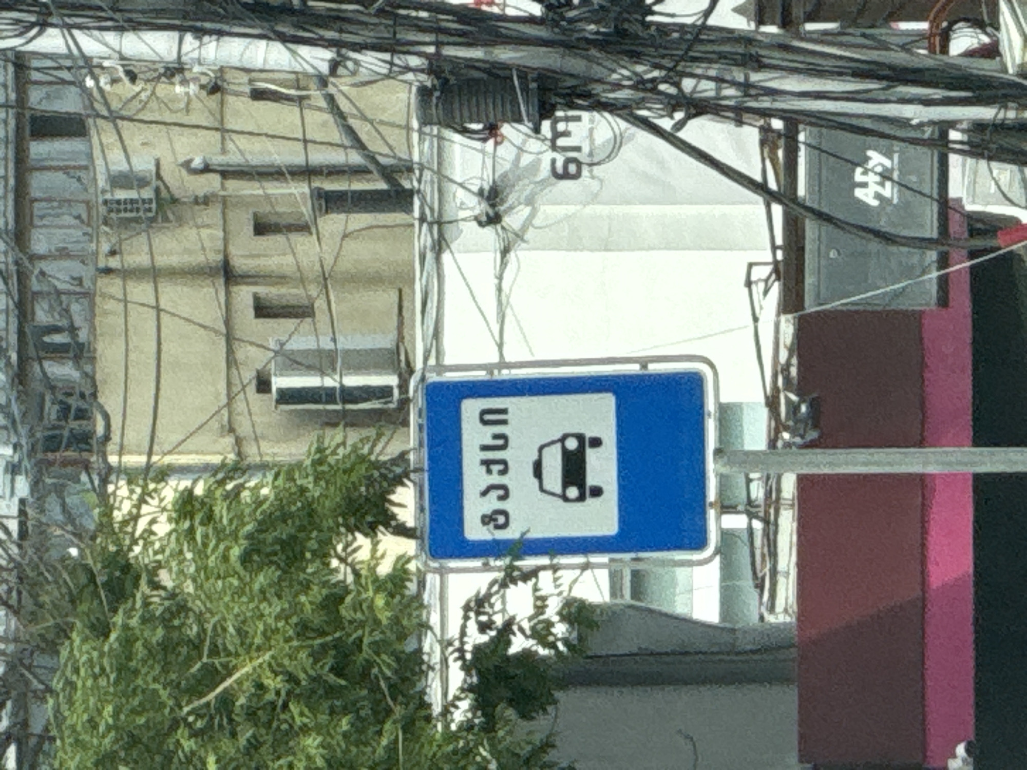 The image shows a blue and white traffic sign with the Georgian word "ტაქსი" (taxi) and a silhouette of a car with a taxi sign on its roof. The sign is positioned on a metal pole, and behind it, there is a building with windows and an air conditioning unit. A tree with green foliage is to the left of the sign. Electrical wires and poles are prominent in the foreground and background. To the right of the sign, another building is visible with the inscription "6M" in white text. Further to the right, a gray electrical box with the white logo "AFY" is mounted. The scene appears to be in an urban environment, likely in Tbilisi, Georgia, given the script on the traffic sign.