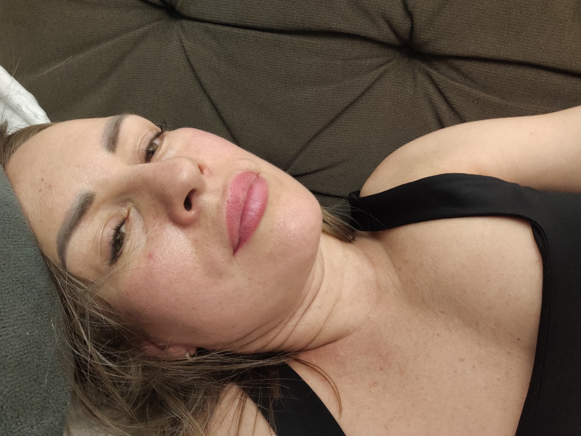 A close-up, high-angle, slightly blurry photograph shows the face of a woman lying on her back on a textured, dark brown sofa. She has blonde hair, fair skin with some blemishes, and is wearing a black top. Her eyes are open and looking towards the camera. Her lips are slightly parted and painted with pink lipstick. The woman appears to be in a relaxed state, with her head tilted to the right. The background is dominated by the sofa, which has a quilted pattern. The lighting is soft and diffused. There are no visible indications of the city of Almaty, Kazakhstan, within the image itself.