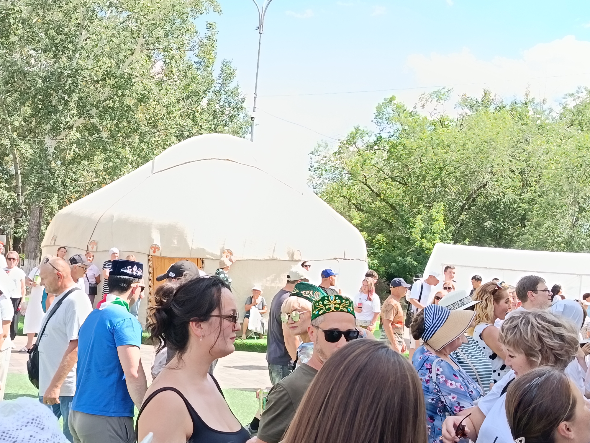 A medium-shot, eye-level, outdoor photograph captures a crowd of people gathered near a large, off-white yurt-like structure, suggesting a cultural event or marketplace. The yurt is positioned in the mid-ground, with its domed roof dominating the left half of the image and extending towards the right. The surrounding environment features lush green trees, indicative of a park or natural setting within the city of Qarağandı, Kazakhstan.
In the foreground, a diverse group of individuals are the primary subjects. On the left, a woman with dark hair and sunglasses is looking to the right, wearing a black tank top. Beside her and slightly behind, a man with a shaved head and sunglasses, wearing a white t-shirt and a black messenger bag, is looking towards the viewer. Further back, a man in a blue t-shirt and a traditional patterned black and white hat is visible. To his right, several other individuals are present, some wearing hats and sunglasses.
The mid-ground reveals more of the yurt and additional attendees. A woman with blonde curly hair, wearing a black and white polka-dot dress, is looking to the right and speaking. She is partially obscuring a woman in a wide-brimmed straw hat with blue and white stripes. Other people, including men and women of various ages, are milling about, some carrying drinks or bags. The yurt itself has an entrance visible, with wooden doors.
The background consists of more trees and a clear, light blue sky with scattered white clouds. The overall impression is a lively gathering, possibly a festival or a community event, taking place on a sunny day in Qarağandı, Kazakhstan. The presence of the yurt suggests a possible theme related to nomadic culture or traditional dwellings. The people are casually dressed, and their interactions appear relaxed, indicating a social atmosphere.