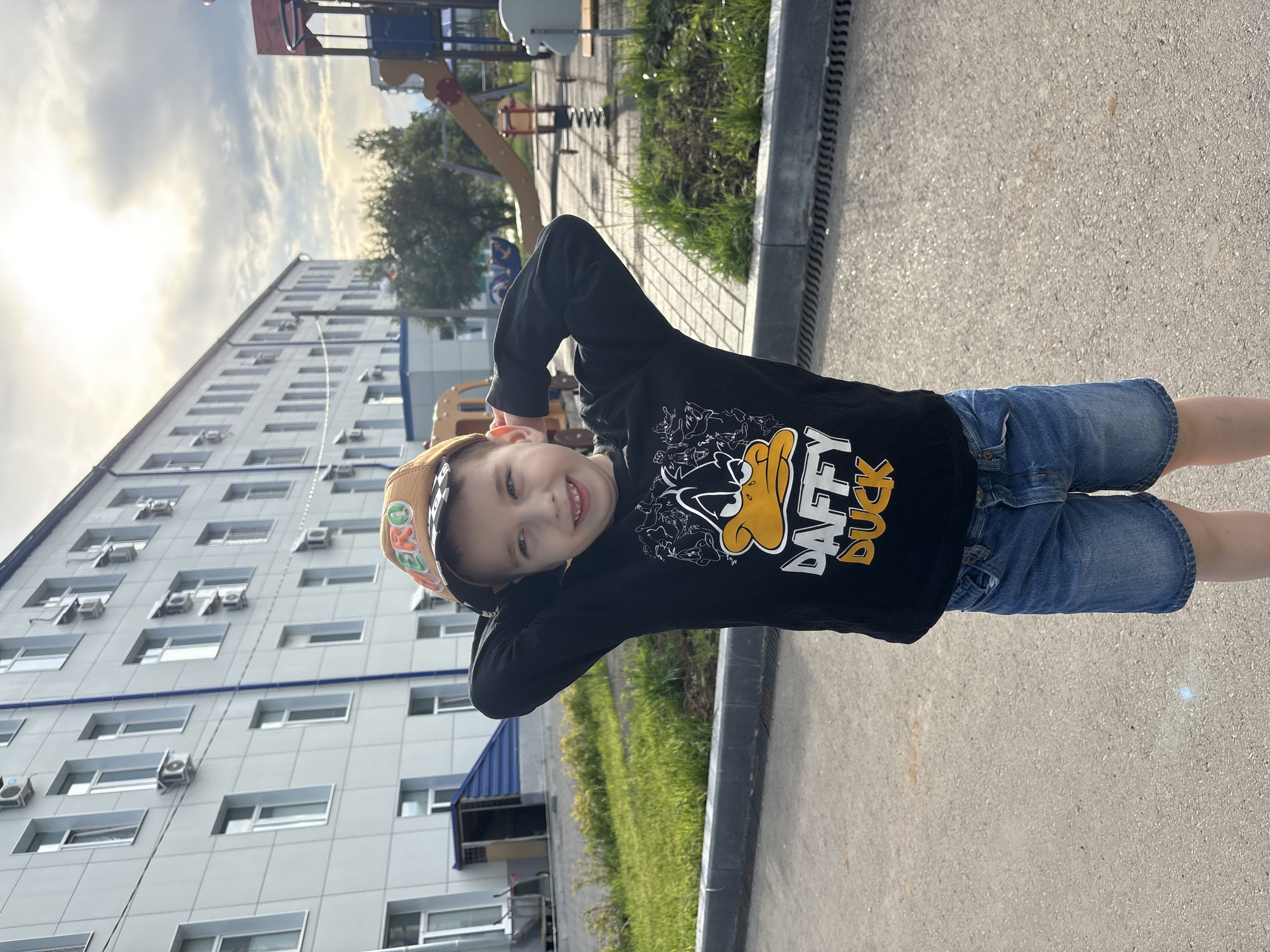 A young boy with blue eyes and light brown hair stands in front of a building, hands behind his head. He wears a black long-sleeved shirt with a Daffy Duck graphic, blue denim shorts, and a brown baseball cap with green lettering. The building appears to be a school or hospital with multiple windows and air conditioning units. In the background, there is a playground with a slide and swings. The scene is set outdoors on a paved area with some grass and trees. The sky is partly cloudy with sunlight visible. The image was taken in Chaikovsky, Russia.