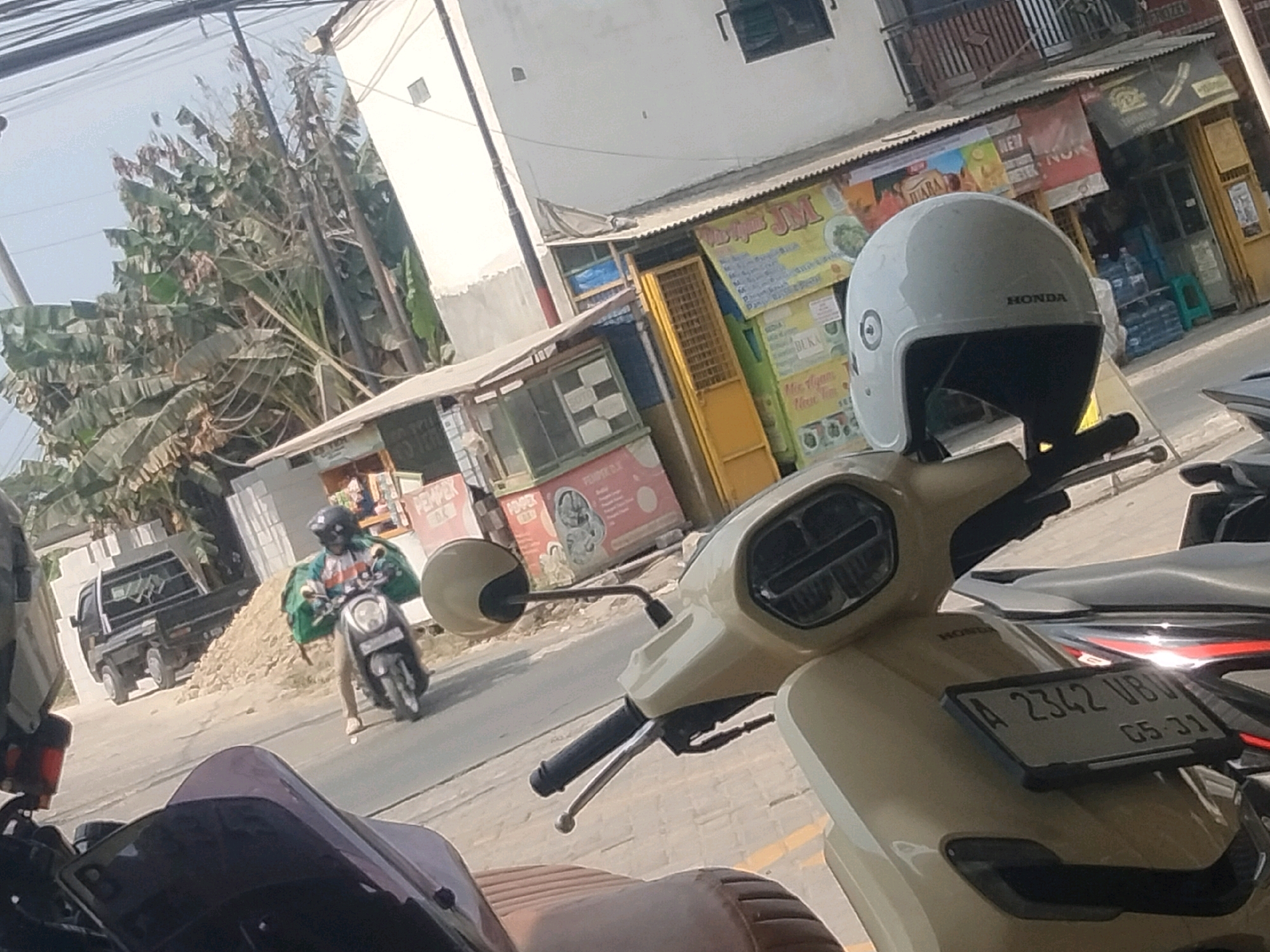 In Pisangan Jaya, Indonesia, a street scene unfolds with several motorcycles as the primary focus. In the foreground, a beige scooter with a black seat is parked, its license plate reading "A 2342 VBU" and below that "05-31". To the left, another motorcycle's front end is visible, with a dark purple windscreen. Further in the background, a person wearing a helmet and a green jacket rides a dark-colored scooter, also with a helmet. Behind this scooter, a small black truck is parked on a dirt embankment. The setting appears to be a street with various small shops and stalls lining the side. Overhead, power lines crisscross the sky, and lush green trees with large leaves, possibly banana trees, are visible to the left. The overall atmosphere is bright and sunny, suggesting daytime.