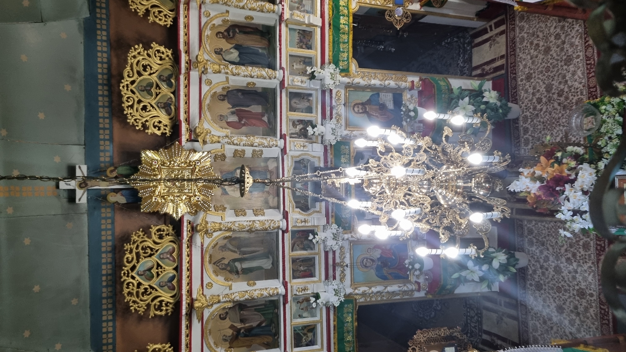 The image shows the interior of an Orthodox church, likely in the city of Vyshka, Ukraine, viewed from above. A large, ornate golden chandelier hangs prominently in the center, illuminated by multiple light bulbs. The walls are decorated with religious icons and gilded ornamental carvings. In the background, a cross with a crucifix is visible, along with more icons depicting religious figures. The floor is covered with a patterned carpet. Floral arrangements are placed on the floor and near the altar area. The overall impression is of a richly decorated and solemn religious space.