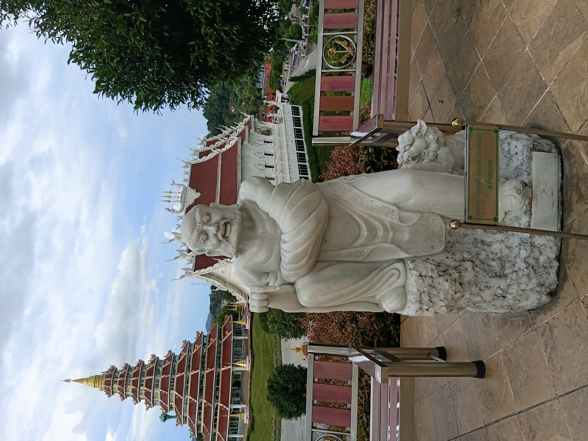 In the foreground, a white marble statue of a seated man with a beard and striking facial features is positioned on a rocky base. To the right of the statue is a smaller, equally white marble statue of a mythical beast, possibly a lion or dragon cub, facing left. A green sign with Thai script is mounted on a pole in front of the statue. Behind the statue, two ornate park benches with stainless steel frames and wooden slats are visible. The benches face away from the viewer, towards a large, multi-tiered, red-roofed stupa with a golden spire in the background. To the right of the stupa, a white building with intricate architectural details and a red roof is partially visible. The scene is set against a backdrop of lush green trees and a cloudy sky. The ground is paved with square tiles in a diamond pattern. The location appears to be a public space, likely a temple or a park, within บ้านเหล่าพัฒนา, Thailand.