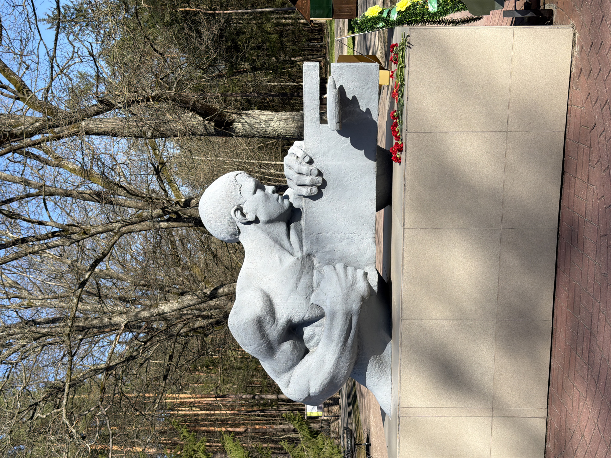 The image shows a gray concrete statue of a man embracing a cross, attached to a beige stone monument. Red flowers are placed at the base of the cross. The statue and monument are situated outdoors, next to a brick pathway. Bare trees are visible in the background, along with a section of a coniferous forest. The location is Камянюцкі сельскі Савет, Belarus.
