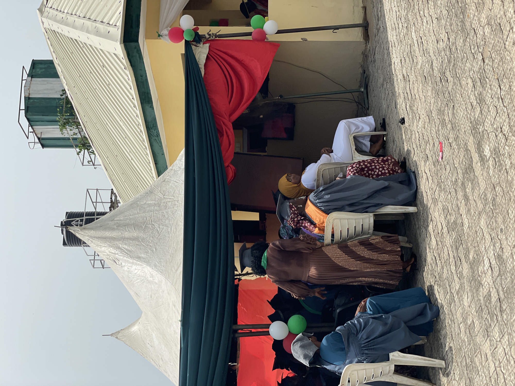 The image depicts an outdoor event in Lokoja, Nigeria. People are seated on white plastic chairs under a large white tent with green and red draping. Red, white, and green balloons are visible, suggesting a celebratory event. A building with a yellow wall and a corrugated metal roof is in the background. A water tank sits on a platform atop the building. Individuals in various attire, including hijabs and traditional clothing, are seated and appear to be attending the event. The ground is paved with textured tiles.