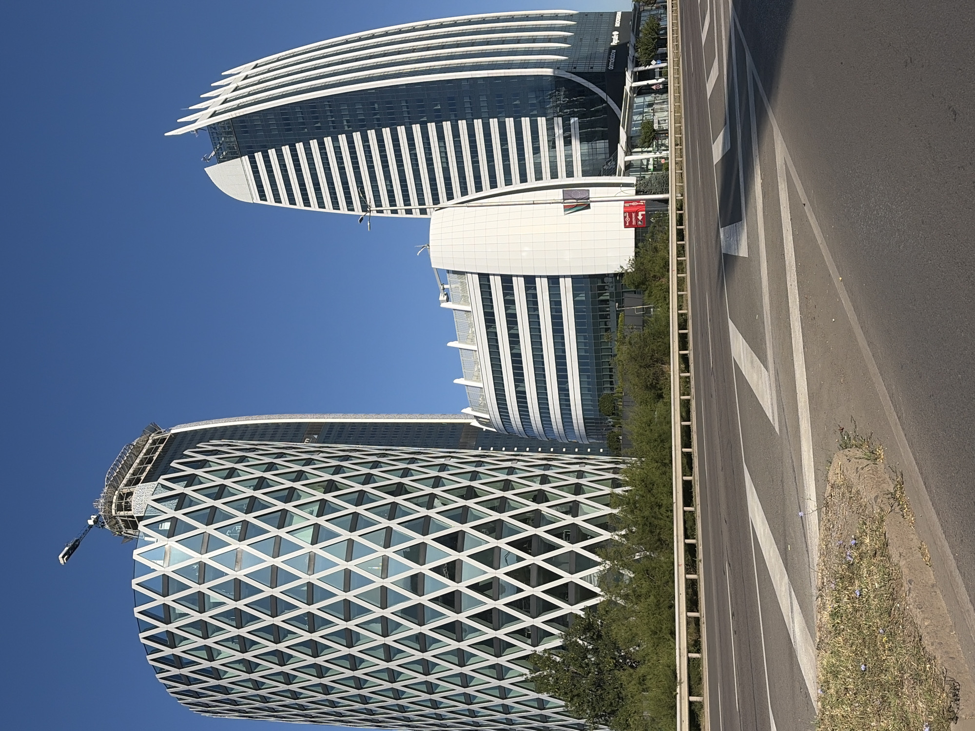 The image shows a street view in Sofia, Bulgaria, featuring a modern architectural complex with several distinct buildings under a clear blue sky. In the foreground, a multi-lane road with white lane markings and pedestrian crossings is visible, bordered by a metal guardrail and a patch of dry grass with a few small purple flowers. Behind the guardrail, lush green bushes and trees line the base of the buildings.
The most prominent structures are three contemporary office buildings. The building on the left is a cylindrical tower with a distinctive white, diamond-patterned facade, giving it a visually striking and geometric appearance. Behind and to the right of this is a taller, partially constructed skyscraper, topped with a crane, indicating ongoing construction. To the far right, a curved, sleek building with a white, fin-like facade dominates the skyline, its reflective glass exterior mirroring the sky. Adjacent to this is a lower, multi-story building with horizontal white banding. Signs are visible at the base of the buildings, including one red sign with a parking symbol and Bulgarian text, and another with logos indicating businesses like "domakibowi" and "Rydes." The overall scene conveys a sense of modern urban development.