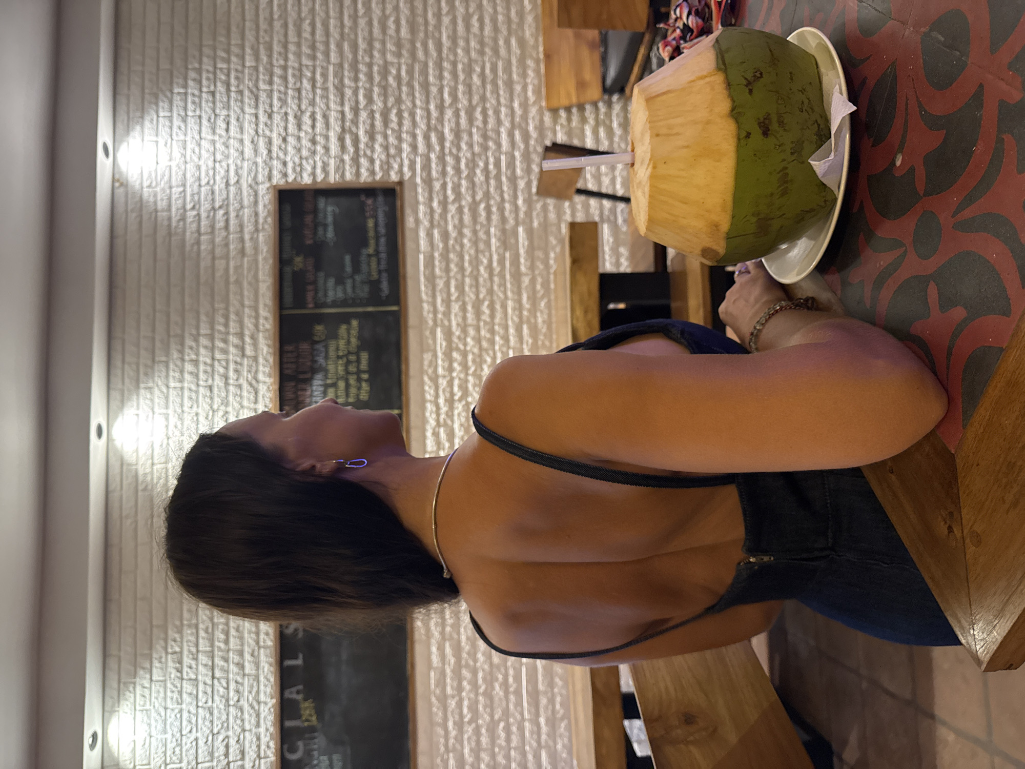 The image shows a person seated at a table in what appears to be a casual restaurant or cafe, viewed from behind and the side. The setting is likely in Kuta Selatan, Indonesia. The person has dark hair, wears a dark-colored top with thin straps and a visible zipper on the side/back, a silver choker necklace, and silver paperclip-shaped earrings. A beaded bracelet is worn on their right wrist.
On the table in front of the person is a large, partially husked green coconut served on a white plate with a straw, indicating it is a drink. The table surface is dark reddish-brown with intricate black patterned designs.
The background features a white brick wall illuminated by several bright, recessed lights, suggesting an evening or nighttime scene. Two dark chalkboards hang on the wall. The left chalkboard clearly displays the word "SPECIALS" at the top, with other, mostly illegible text below it. The right chalkboard appears to list menu items, with categories such as "DRINKS," "SMOOTHIES," "COFFEE," "TEA," and "JUICES" faintly visible, along with some specific, indistinct beverage names like "Traditional" and "Ginger." The overall atmosphere is relaxed and informal.
