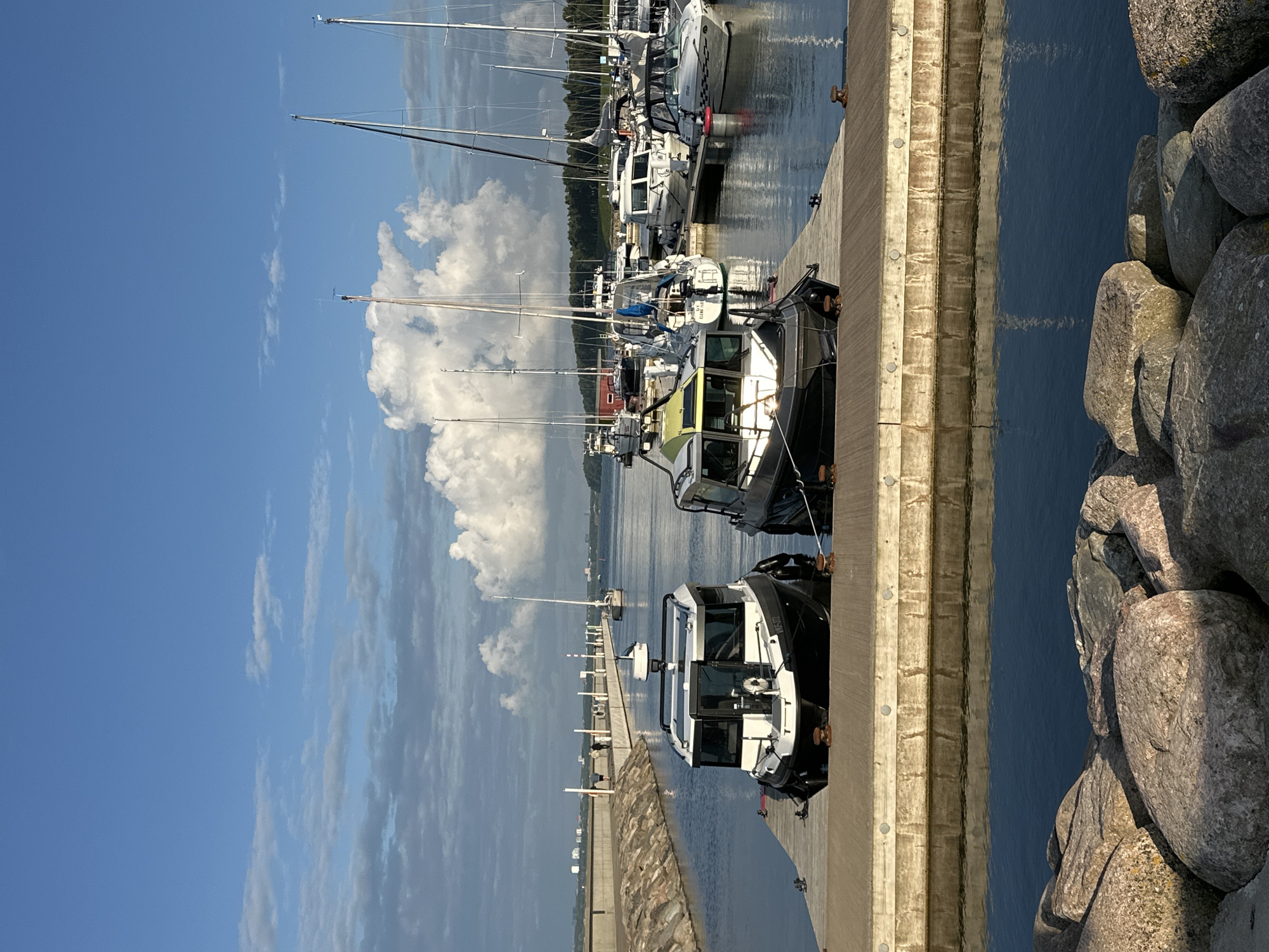 In Tallinn, Estonia, two motorboats are docked side-by-side on a wooden pier in a marina. The boat on the left is predominantly white with a black hull and a radar dome on its roof. The boat on the right has a black hull and a white cabin with green accents. Beyond the two boats, numerous sailboats are moored, their masts reaching towards a partly cloudy blue sky. In the background, a concrete breakwater extends into the water, with a few people walking along its top. The water is calm, reflecting the sky and the boats. Large rocks are visible in the foreground of the image.