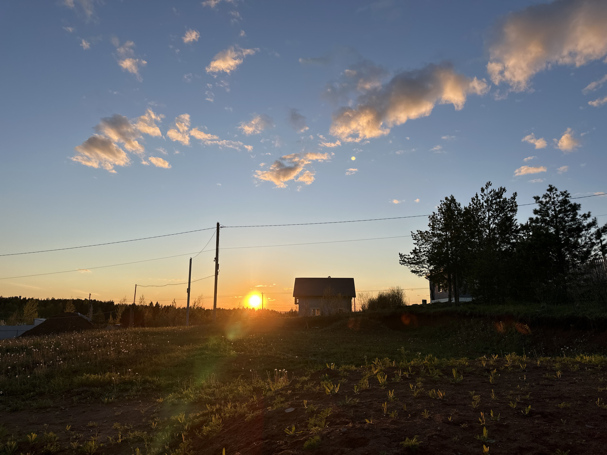 The image depicts a rural scene near Прикамский, Russia at sunset. A small house is positioned between the viewer and the setting sun. A utility pole stands to the left of the house, with a power line extending across the frame. The foreground consists of a field with sparse vegetation. To the right of the house are trees. The sky is a mix of blue and orange, with scattered clouds.