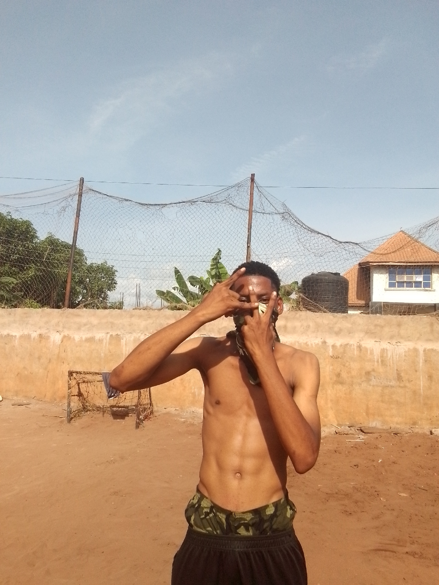 A shirtless dark-skinned man is posing in an outdoor area in the city of Unknown, Unknown. He is wearing camouflage shorts with black pants over them. The man is holding his hands up to his face, partially obscuring it with his fingers. A black bandana is covering his nose and mouth. In the background, there is a low wall, a soccer goal, a dilapidated net fence, trees, a water tank, and a house with an orange roof. The ground is reddish-brown, and the sky is blue.