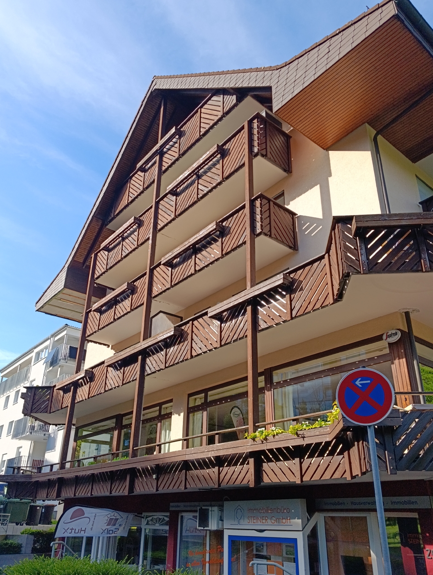 The image shows a four-story building with prominent wooden balconies and a gabled roof. The building's facade is primarily cream-colored, with dark brown wooden supports and balcony railings. A "no stopping" traffic sign is visible in the foreground, partially obscuring the lower part of the building. The street level of the building features businesses, including one labeled "Immobilienbüro - STEINER GmbH." Another building, white with balconies, is visible across the street. The sky is blue and clear. The specific location within an unidentified city is unknown.