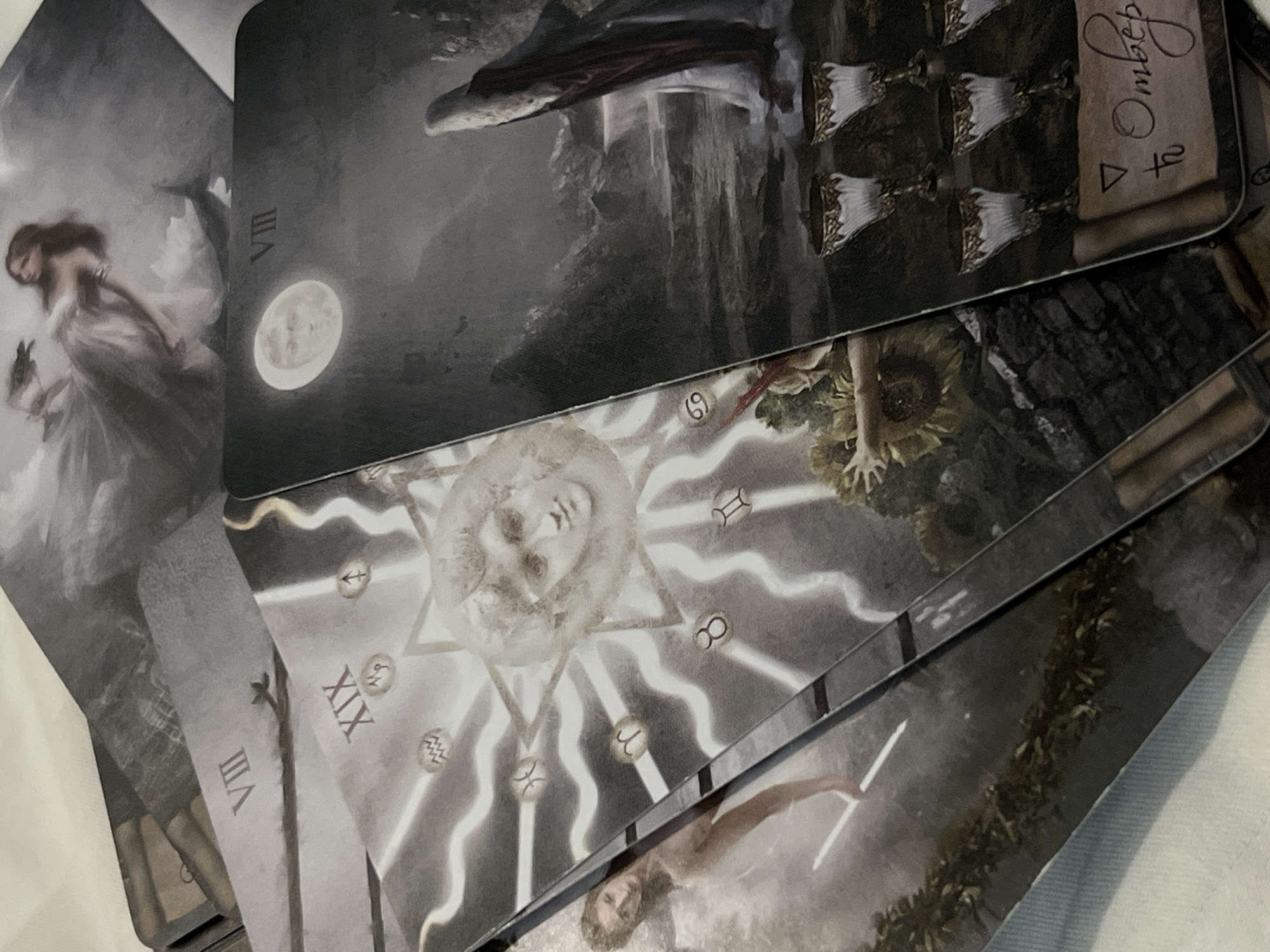 The image shows a close-up view of several tarot cards laid out on a light-colored surface, possibly fabric. The cards are overlapping and are primarily in grayscale or muted tones. The visible cards include:
* One card showing a woman in a flowing dress.
* A card with "VIII" and an image of a moon.
* A card with the Roman numeral "XIX"
* A card featuring an image of the sun with astrological symbols.
* A card depicting cups.
There is no indication of the location being in Bac Nha Trang Ward, Vietnam, within the provided image. The objects and their arrangement are limited to the tarot cards themselves and the surface they rest on. There are no discernible actions or interactions captured within the frame.
