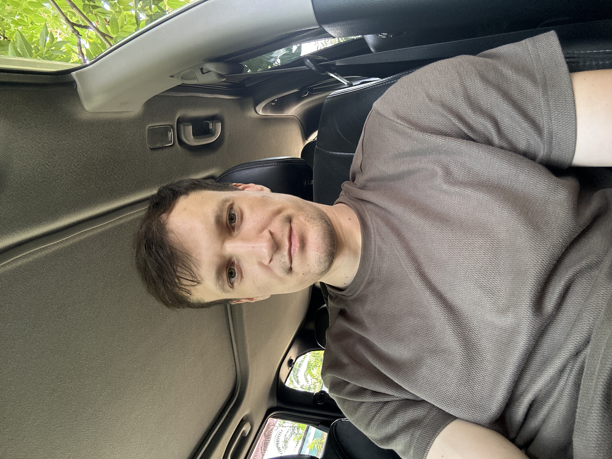 A man is sitting in the driver's seat of a car, looking at the camera. He is wearing a brown t-shirt. The car's sunroof is open, and leafy trees are visible outside. The image appears to have been taken during the day in Makiv, Ukraine.