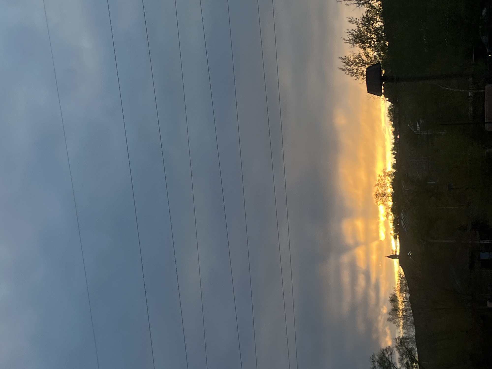 The image captures a sunset scene in an unidentified area possibly near the city of Krasnoyarsk, Russia, judging by the electrical pylons commonly seen in Russian infrastructure.
In the foreground, a series of parallel electrical wires stretch across the sky. The sky itself is a mix of blue and grey clouds with a pronounced area of intense orange sunlight breaking through. On the right side, silhouettes of trees and structures. The golden light highlights what appear to be tree branches and some form of building or utility structure.
