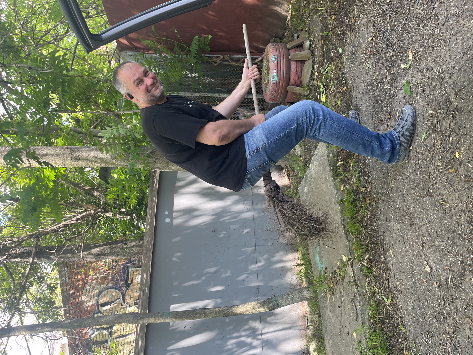 A man is depicted outdoors, horizontally oriented as if mid-air, playfully posing on a broomstick. He is middle-aged, with a beard and graying hair, wearing a black t-shirt, blue jeans, and gray athletic shoes, with an earbud in his left ear. He smiles broadly at the camera, holding a wooden stick with a bundle of bristles at one end, positioned between his legs to mimic a witch's broom.
The scene is set in Krasnodar, Russia, under bright daylight with clear shadows. In the background to the left, there are lush green trees and foliage, a brick wall partially covered with abstract graffiti, and a gray concrete or metal wall. To the right, a red-painted corrugated metal structure or shed is visible, along with a fence and two red-painted tires stacked and used as planters with small green plants. The ground beneath consists of dirt, gravel, and patches of grass, with a concrete slab visible. A dark car door is partially seen in the upper part of the frame, possibly serving as a prop for the illusion of flight. No discernible text is visible on clothing or graffiti.