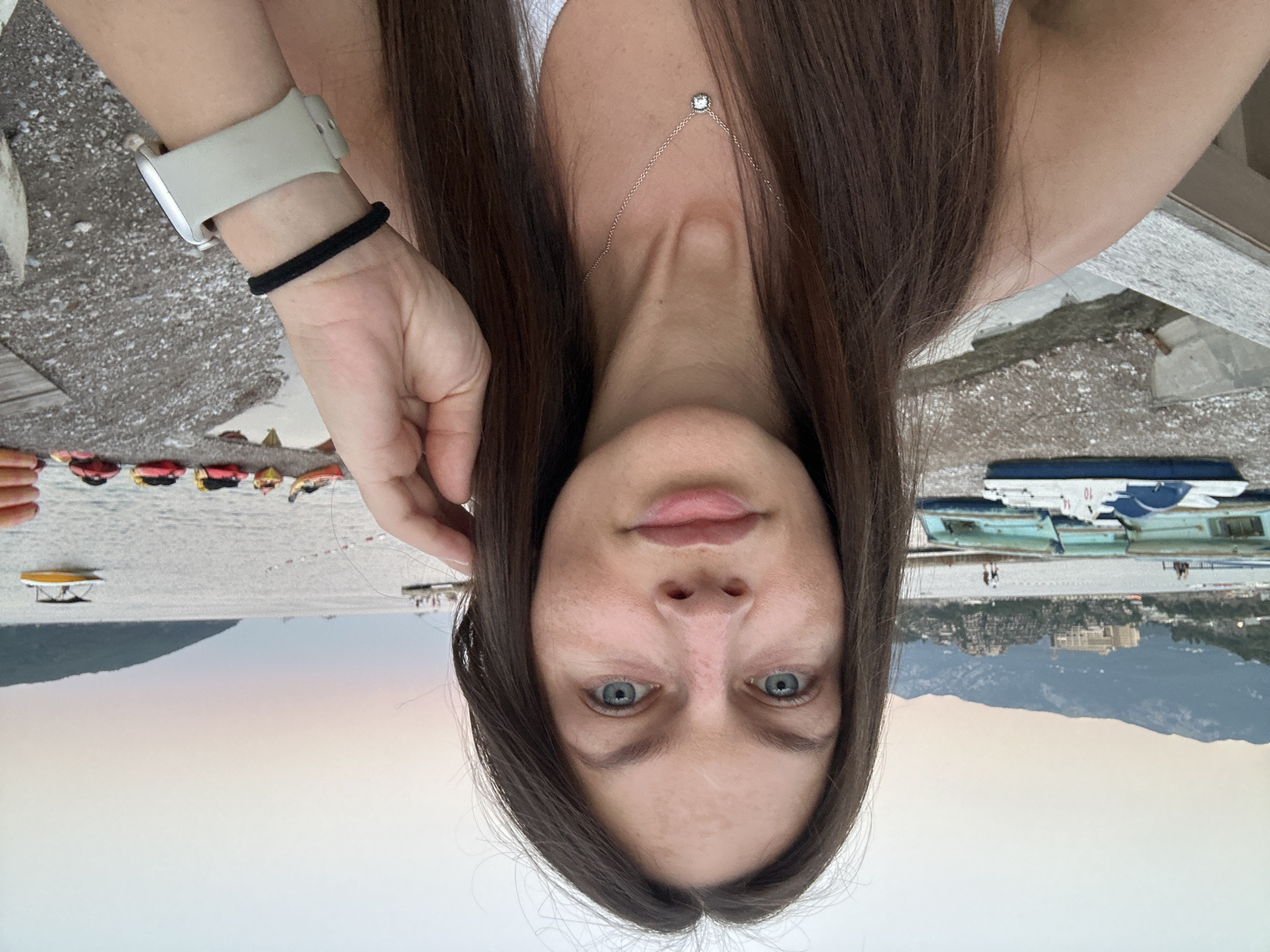 The image is a selfie taken by a woman with long, dark brown hair and blue eyes, in Budva, Montenegro. She is wearing a white top and a necklace with a small pendant. On her left wrist, she wears a smartwatch with a light-colored band and a black hair tie. She is touching her hair with her right hand. The background shows a beach with several kayaks of various colors lined up on the shore. In the water, there are pedal boats and a small yellow boat. Behind the beach, there are hills and mountains, and buildings are visible on the slopes. The sky is overcast with soft pastel colors, suggesting it is either dawn or dusk. The overall setting appears to be a coastal resort area.