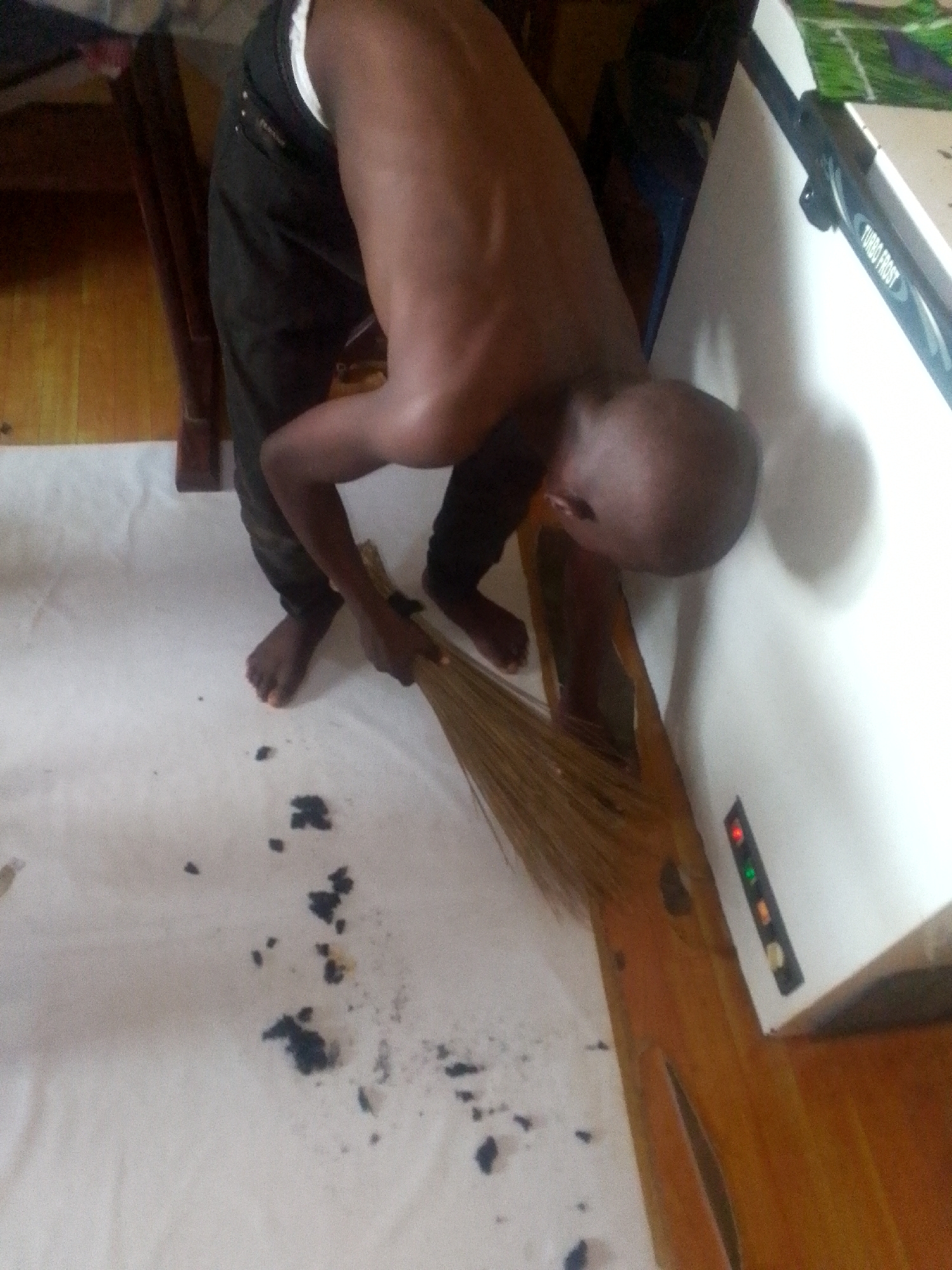 An individual with dark skin, shirtless and wearing dark trousers, is captured in an indoor setting, bent over and engaged in sweeping. The person, who is barefoot, is using a traditional broom made of natural fibers to clear dark, scattered debris from a light-colored floor or sheet. To the right of the person, a white appliance or piece of furniture is visible, featuring a label that reads "TURBO FROST." This white object also displays a vertical panel with illuminated indicator lights, showing red, green, and amber colors. Various wooden elements are present in the background and foreground, suggesting the room's furnishings or structure. The scene depicts a routine cleaning activity within what appears to be a domestic space.