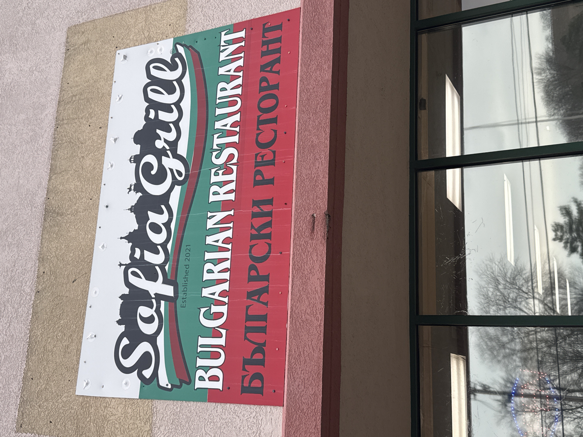 The image shows the exterior of the Sofia Grill restaurant in Des Plaines, Illinois, USA. The sign displays "Sofia Grill" in stylized lettering above an outline of a cityscape. Below this is "Bulgarian Restaurant" in English and Bulgarian. The sign is mounted on a building. Adjacent to the sign is a window reflecting the outside environment.
