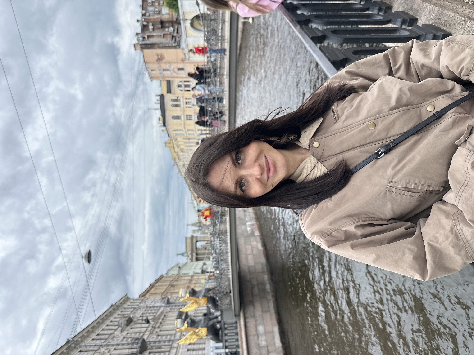 In Saint Petersburg, Russia, a young woman with long, dark hair stands on a bridge overlooking a canal. She is wearing a tan jacket with a corduroy collar and a black crossbody bag. Her gaze is directed towards the camera with a slight smile. In the background, the canal is bordered by buildings, and a bridge adorned with golden griffins is visible. People are walking across the bridge, and the sky is overcast.