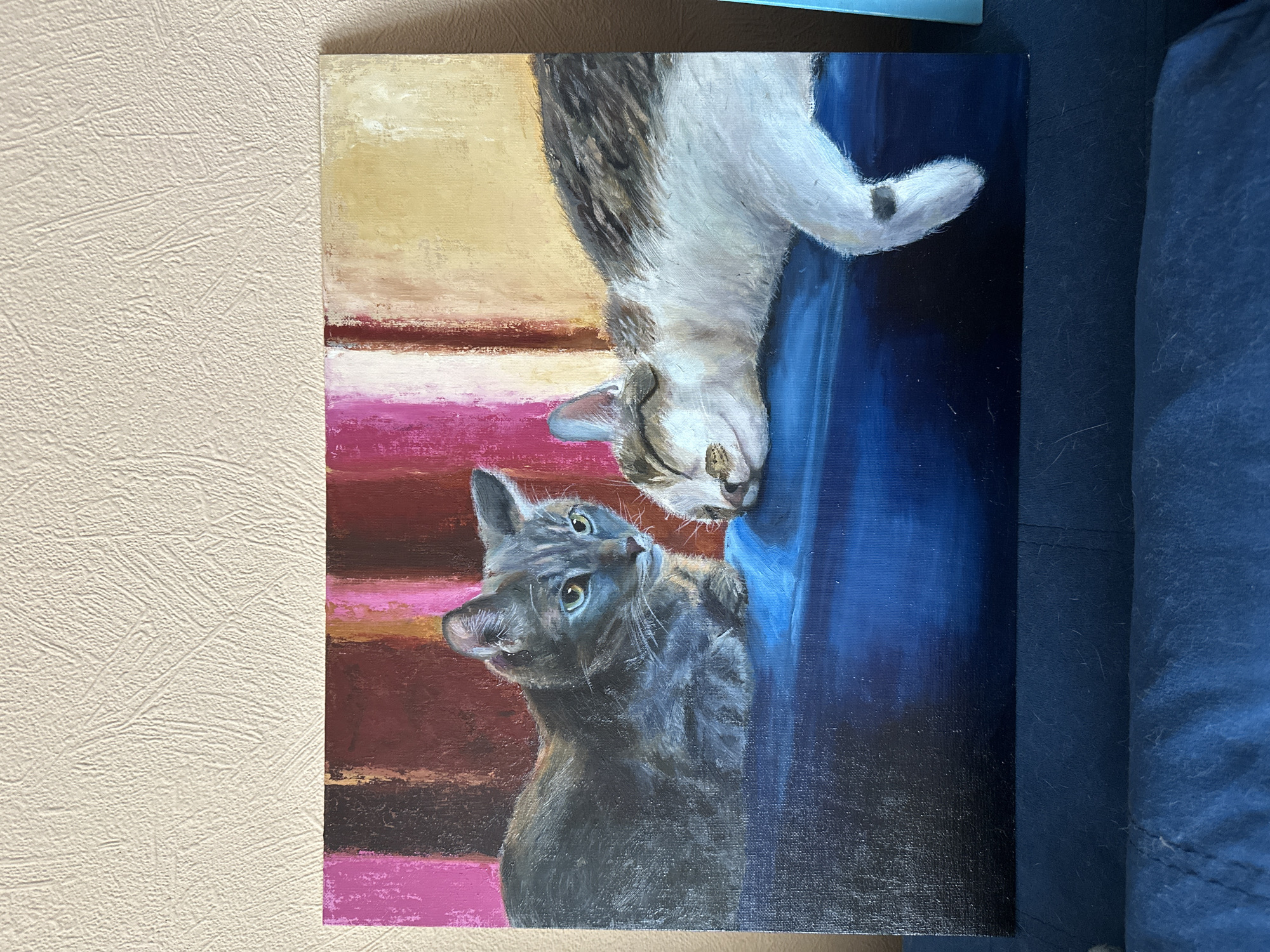 The image depicts an oil painting on canvas, propped against a beige textured wall and a blue fabric surface. The painting features two cats in an unusual, upside-down composition. The upper cat is a gray tabby with white markings, depicted upside-down, nose near the bottom cat's head. The lower cat is a gray cat looking upwards, its green eyes gazing at the tabby. The background consists of horizontal bands of varying colors - a beige/yellow at the top, followed by pink, brown, and pink again. A dark blue area, possibly representing a couch or similar object, is behind the cats. There are no other people, subjects, or actions depicted beyond the cats' interaction. There is no evidence in the image to suggest a location in Krasnoyarsk, Russia.