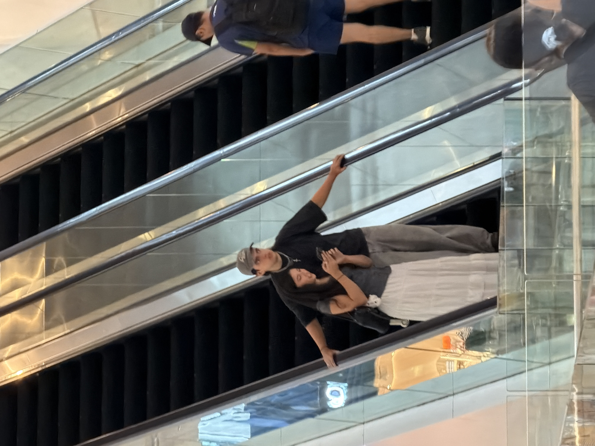 A young couple stands on an escalator in what appears to be a shopping mall. The woman, wearing a white tiered skirt and a dark grey top, holds a mobile phone and is looking down at it. The man, in grey pants and a dark t-shirt with a baseball cap, has his left arm extended and rests his hand on the escalator railing. A man wearing a blue t-shirt and blue shorts with a backpack is ascending the escalator in the background. The scene is captured from a slightly elevated perspective, looking down at the escalators. The environment is brightly lit with reflective surfaces typical of an indoor commercial space. The location is Yuzhong District, China.