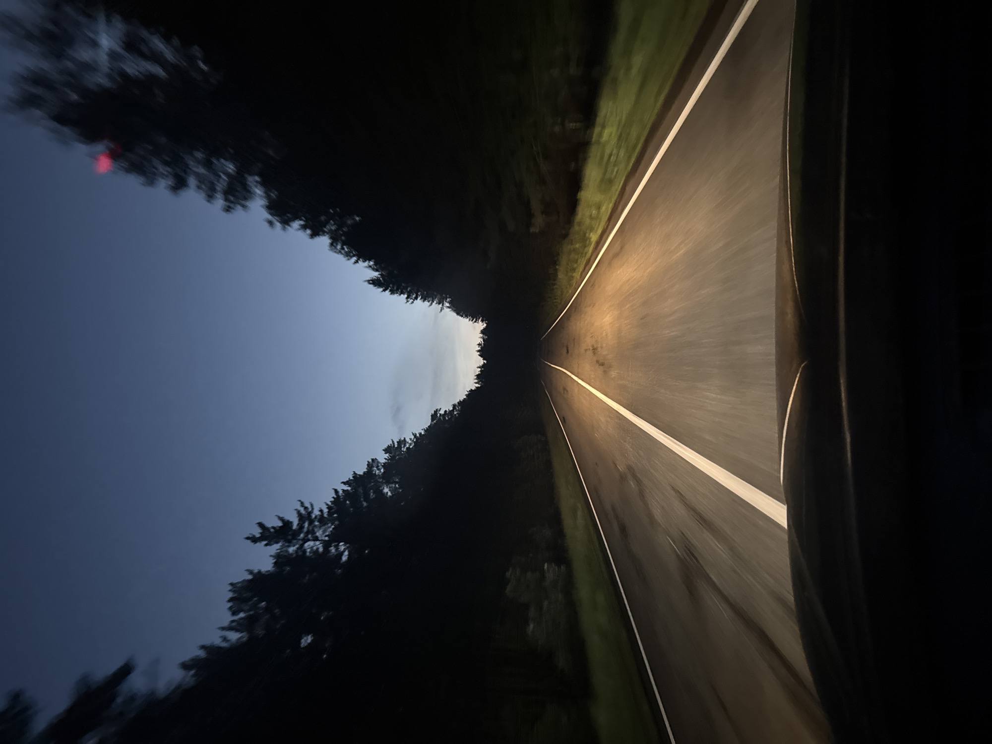 The image depicts a view from the driver's perspective, looking forward along a dark asphalt road through a forest. The sky above is a deep twilight blue, with a lighter gradient near the horizon. The road has a white line marking the center and white lines on the edges. Headlights from the vehicle illuminate the road ahead, casting a warm glow. Tall, dark pine trees line both sides of the road, creating a sense of enclosure. The overall impression is that of driving on a rural road at dusk or early night. The location, Балаки, Russia, is a rural area, consistent with the visual cues of a forest road. There are no people or other vehicles visible in the image. The image is a photograph taken from within a moving vehicle.