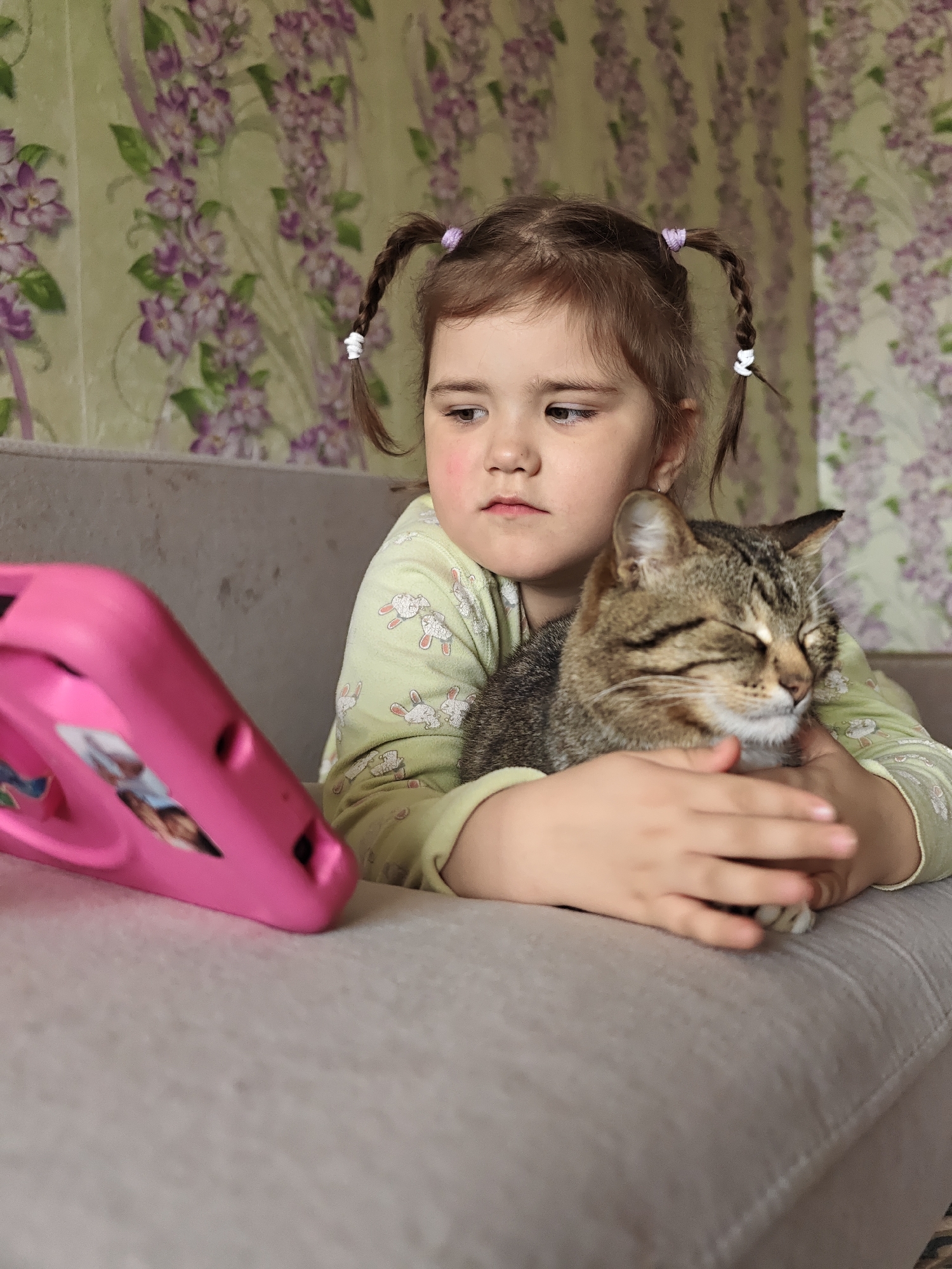 A young girl with brown hair styled in pigtails held with purple and white bands sits on a gray couch, embracing a tabby cat with closed eyes. The girl wears a light green pajama top with a pattern of white rabbits. A bright pink tablet stands in the foreground to the left of the girl. The background features a wall with floral wallpaper depicting purple and green flowers. This photo was likely taken indoors in Уральское, Russia.