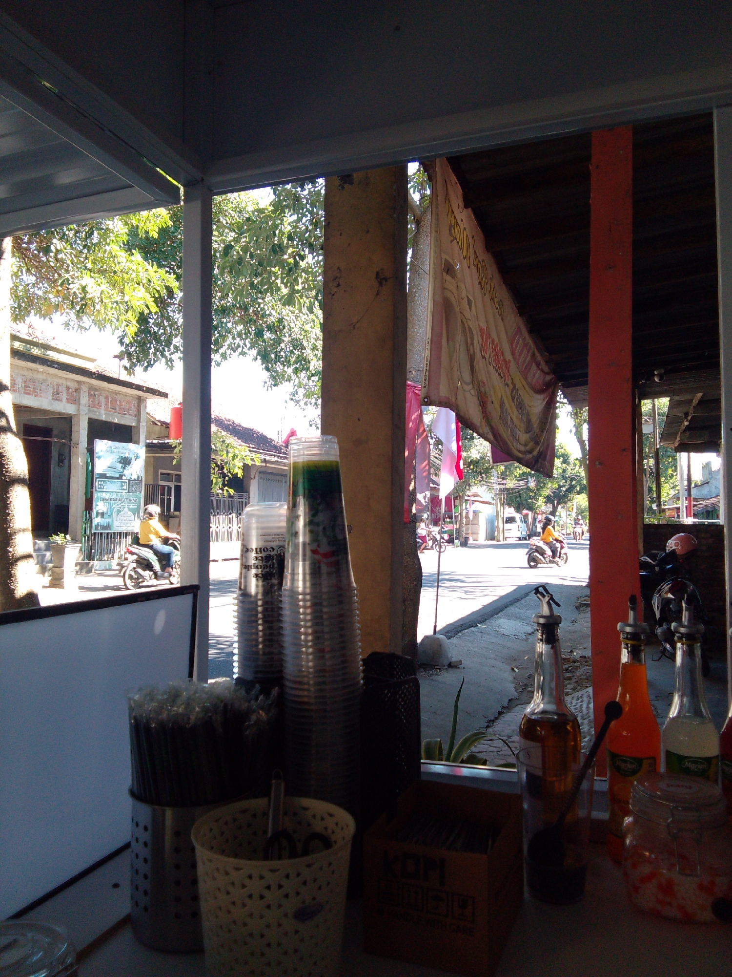 This image captures a street-level view of a small food or drink stall in Trenggalek, Indonesia. The foreground features a display of items for sale or use, including stacks of clear plastic cups, a metal container filled with straws, and a white woven basket holding utensils. Several bottles of liquids, possibly syrups or sauces, are lined up, with one bottle containing a dark brown liquid and a spoon. A glass jar filled with colorful, small candies or toppings sits to the right.
Beyond the stall, the scene opens to a street with moderate traffic. A person wearing a yellow shirt and helmet is riding a motorcycle. Buildings with traditional Indonesian architecture, including pitched roofs and open verandas, are visible along the street. Lush green trees provide shade. A large banner, possibly advertising a product or event, hangs across the upper portion of the scene, displaying text that appears to be in Indonesian. The overall impression is of a vibrant, everyday scene in a local Indonesian community.